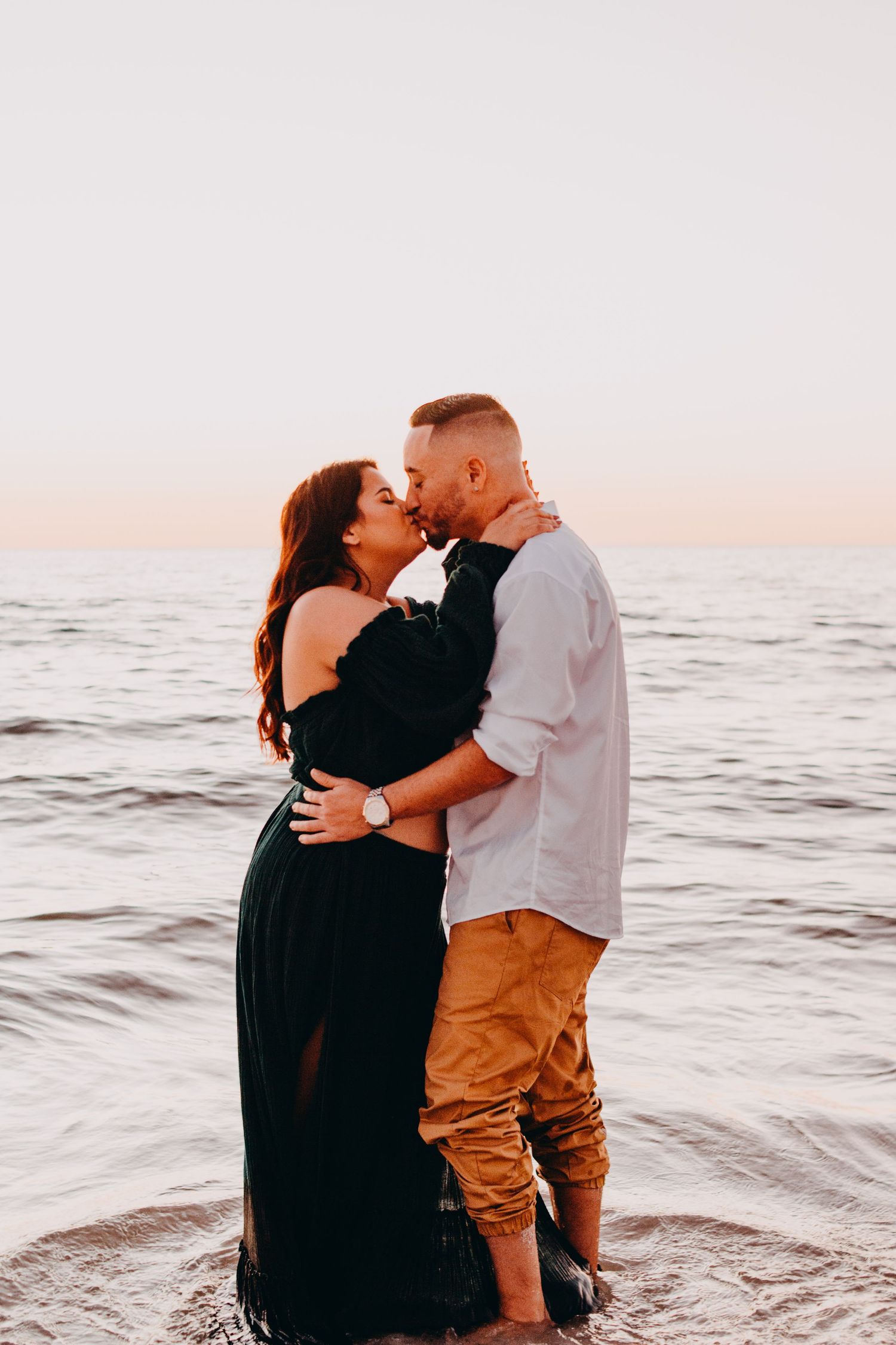 A couple shares a romantic embrace on the beach at sunset with ocean waves in the background.