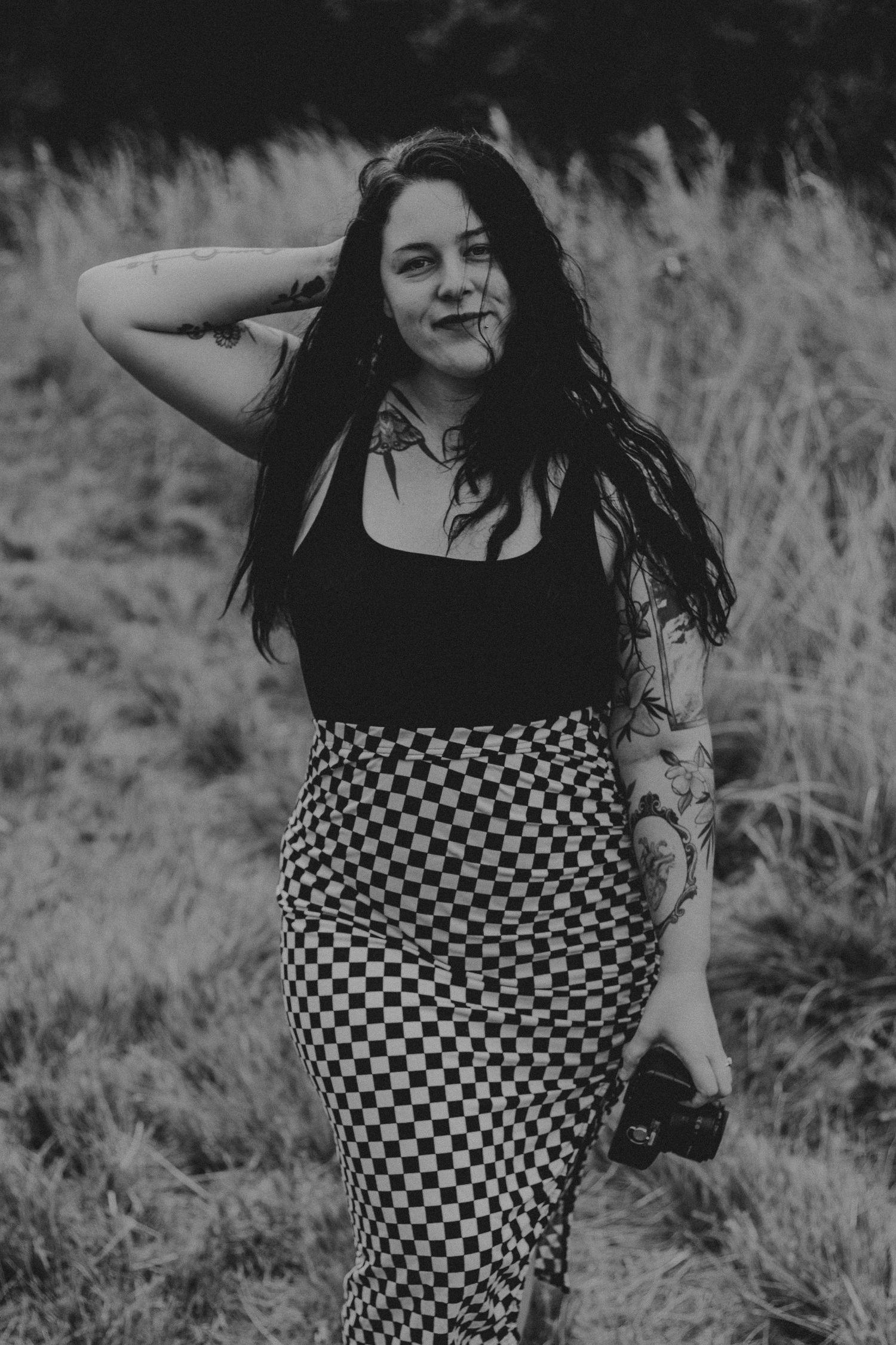 Black and white portrait of someone in a black tank top and checkered skirt standing in tall grass.