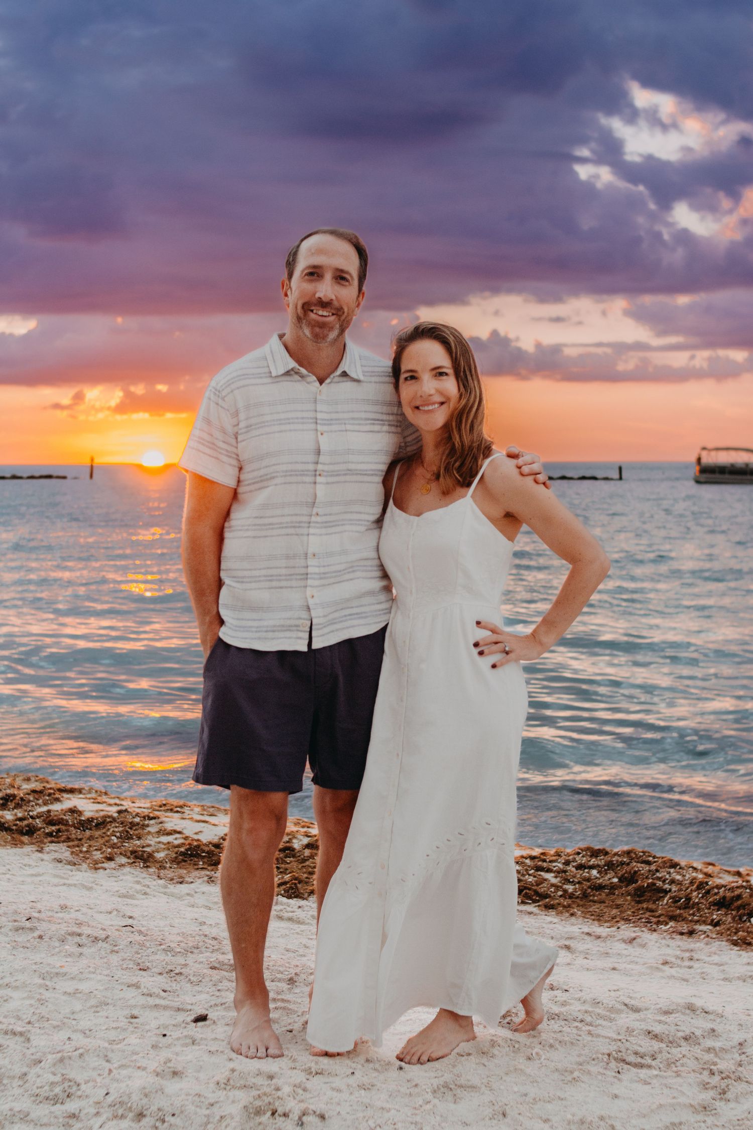 Couple enjoying a romantic sunset photoshoot on a tropical beach with dramatic purple and orange sky.