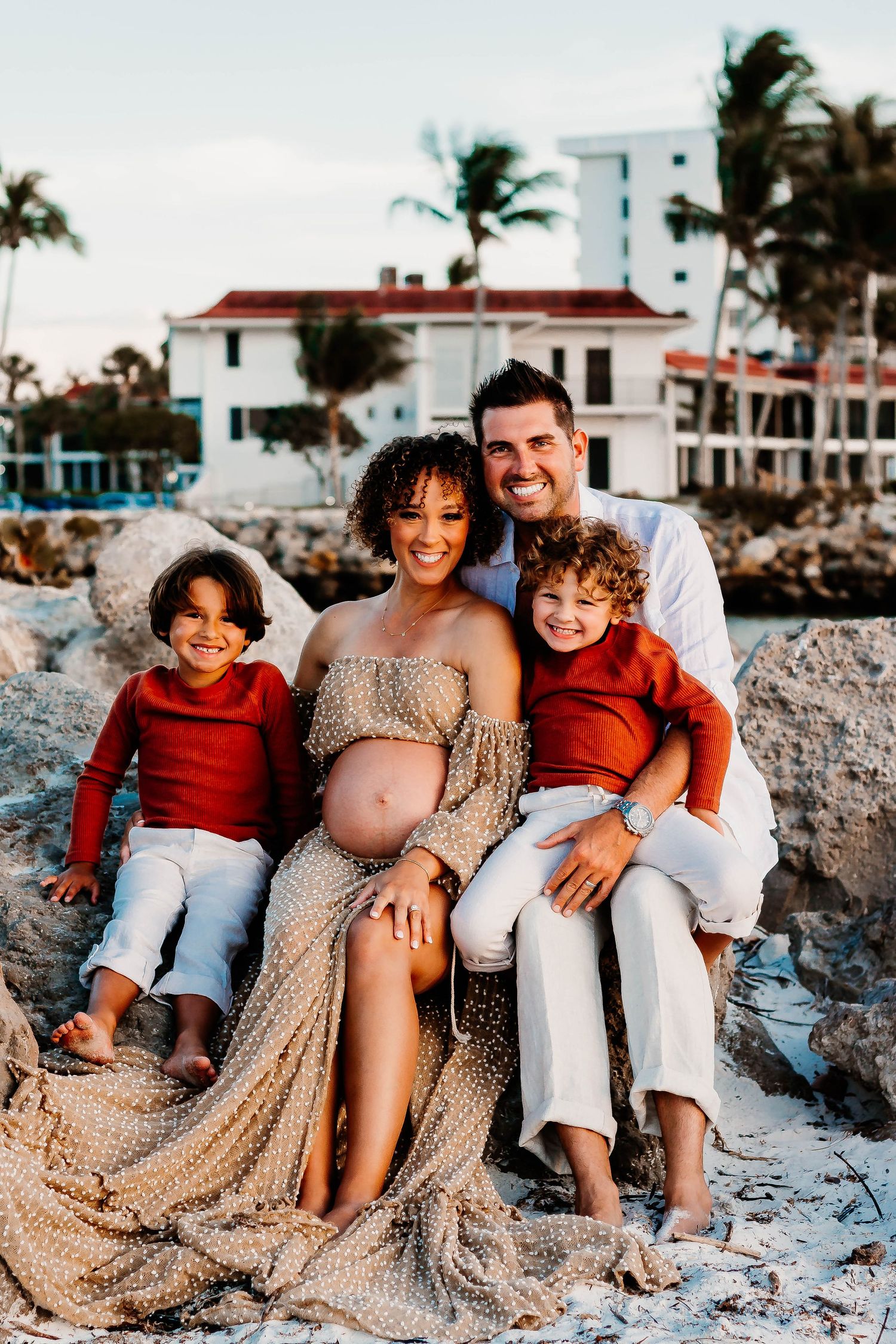 Family wearing red and beige outfits sits together on sandy beach at sunset for casual outdoor photoshoot.