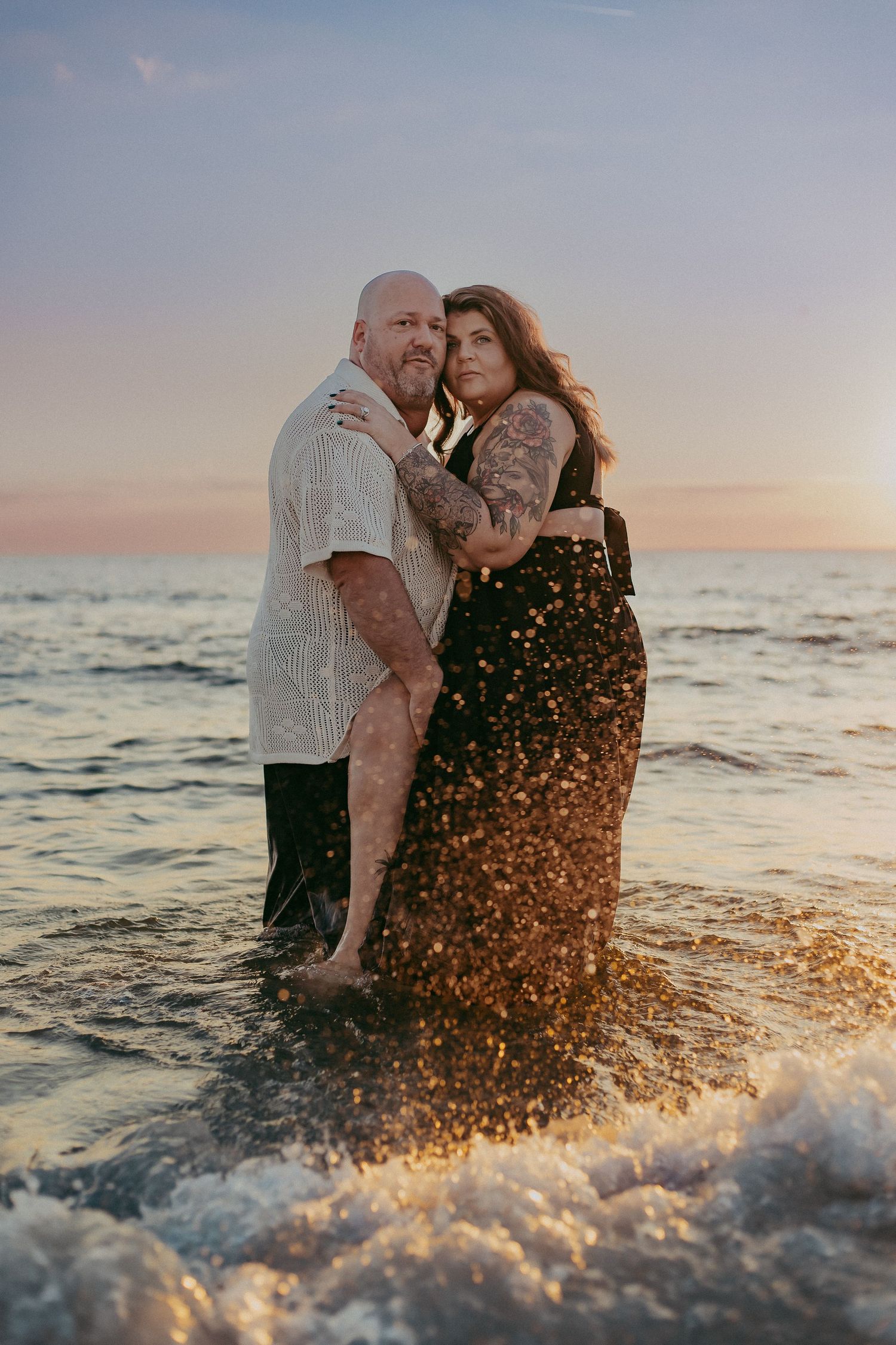 Romantic couple embraces in ocean waves at sunset while water splashes around them creating magical golden light.