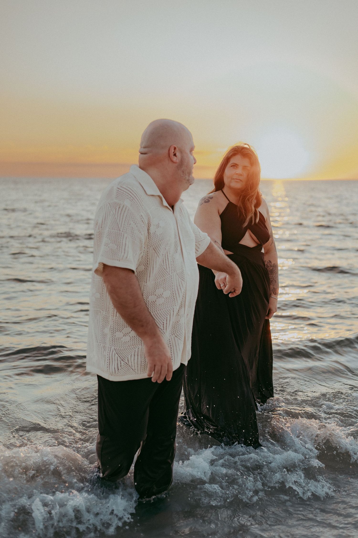 Two people share a romantic moment while standing in ocean waves at sunset, creating a dreamy silhouette against the water.