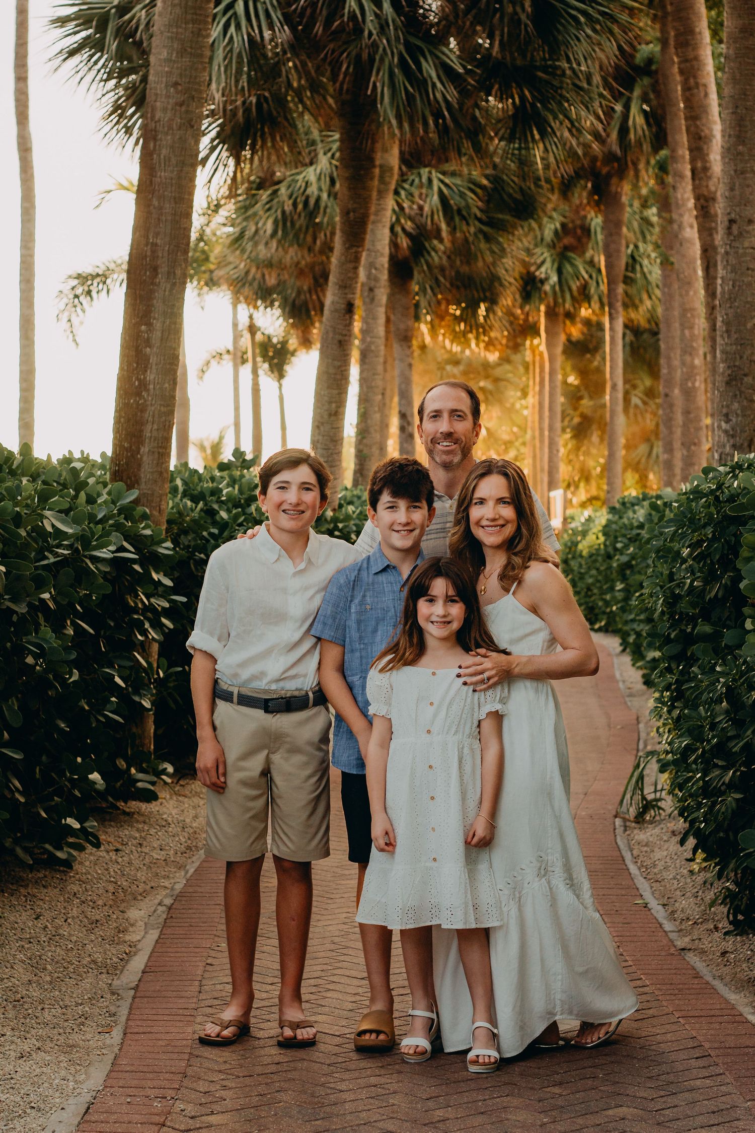 Family portrait series taken on a palm tree-lined brick path at sunset with coordinated neutral outfits.