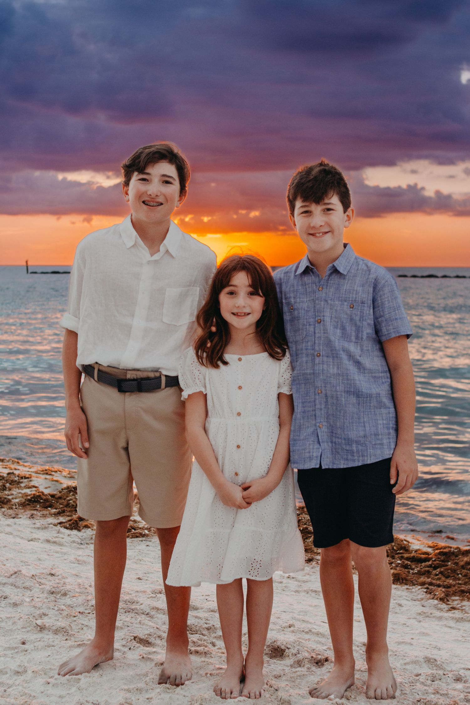 A family stands together on a beach at sunset with dramatic orange and purple sky over the ocean.