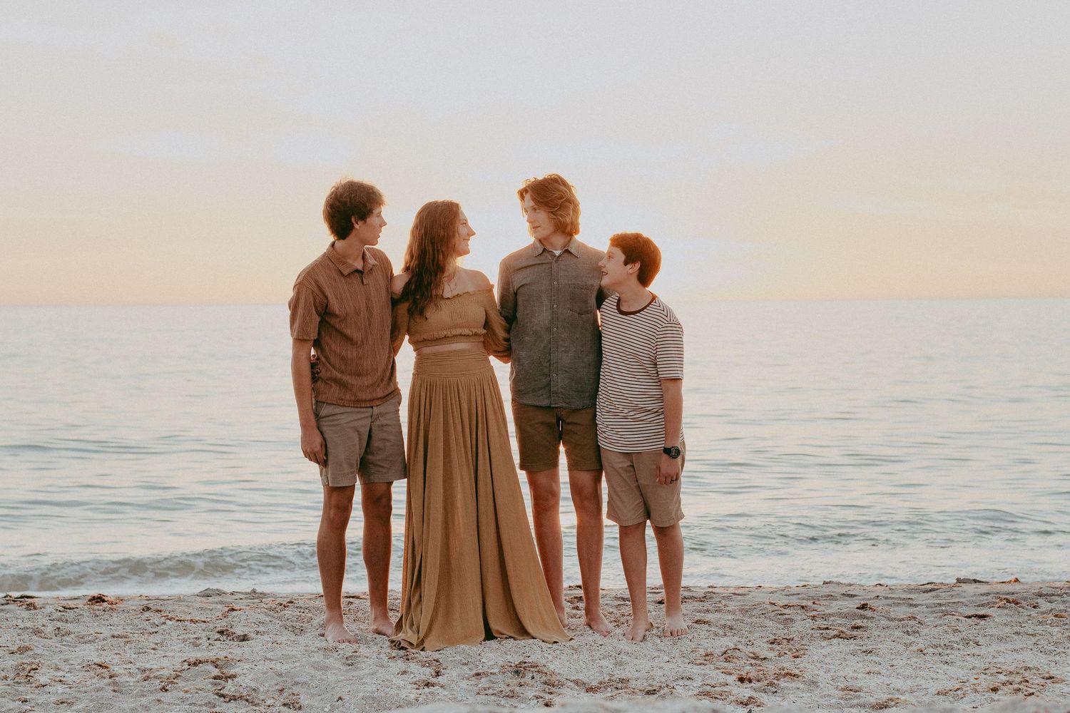A group of people stand together on a sandy beach at sunset looking out at the ocean.