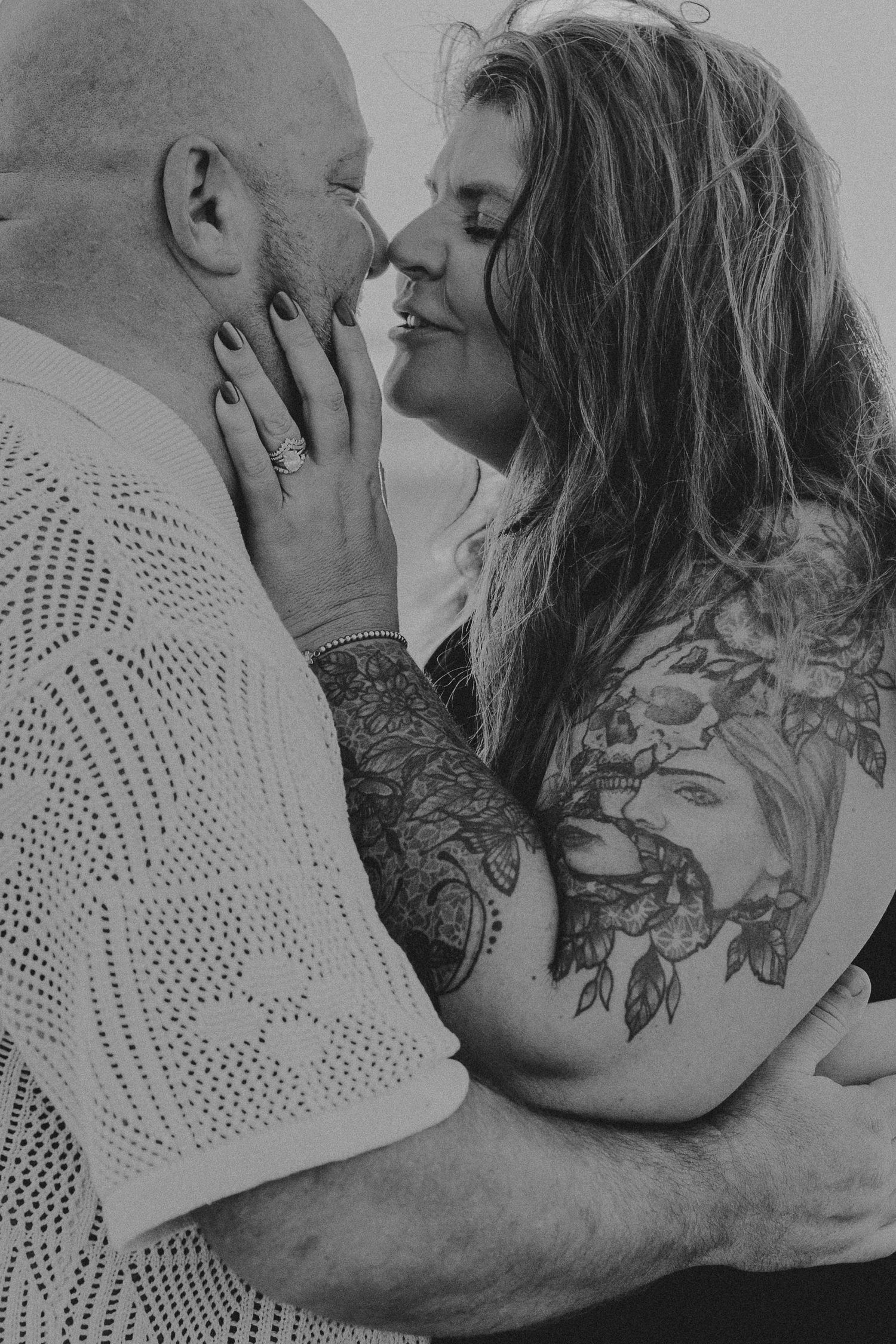 Black and white intimate photo of a couple embracing, with visible tattoos and tender expressions.