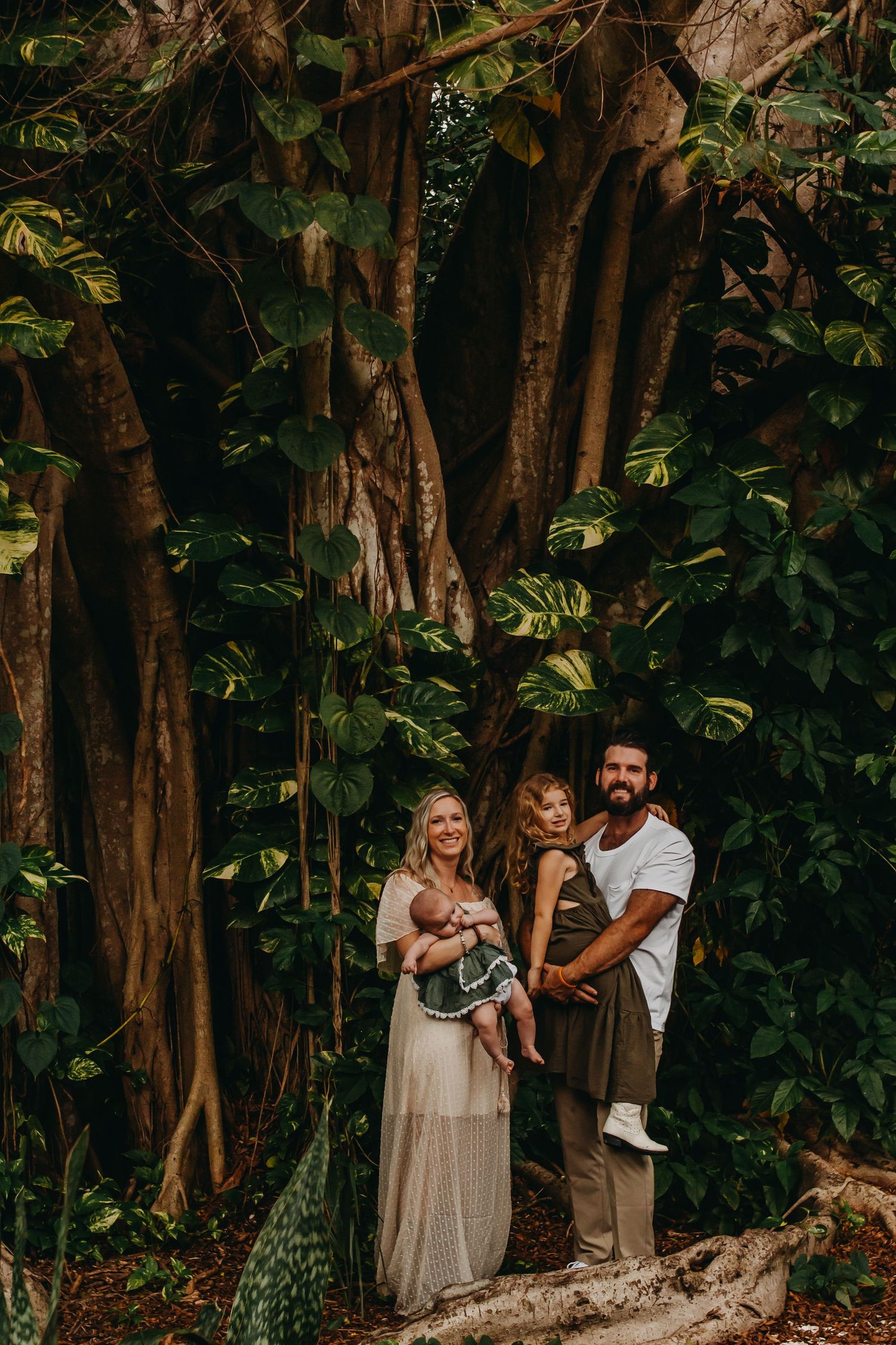 Family of three poses before a massive banyan tree in a lush tropical forest, mother in flowing dress holding newborn.
