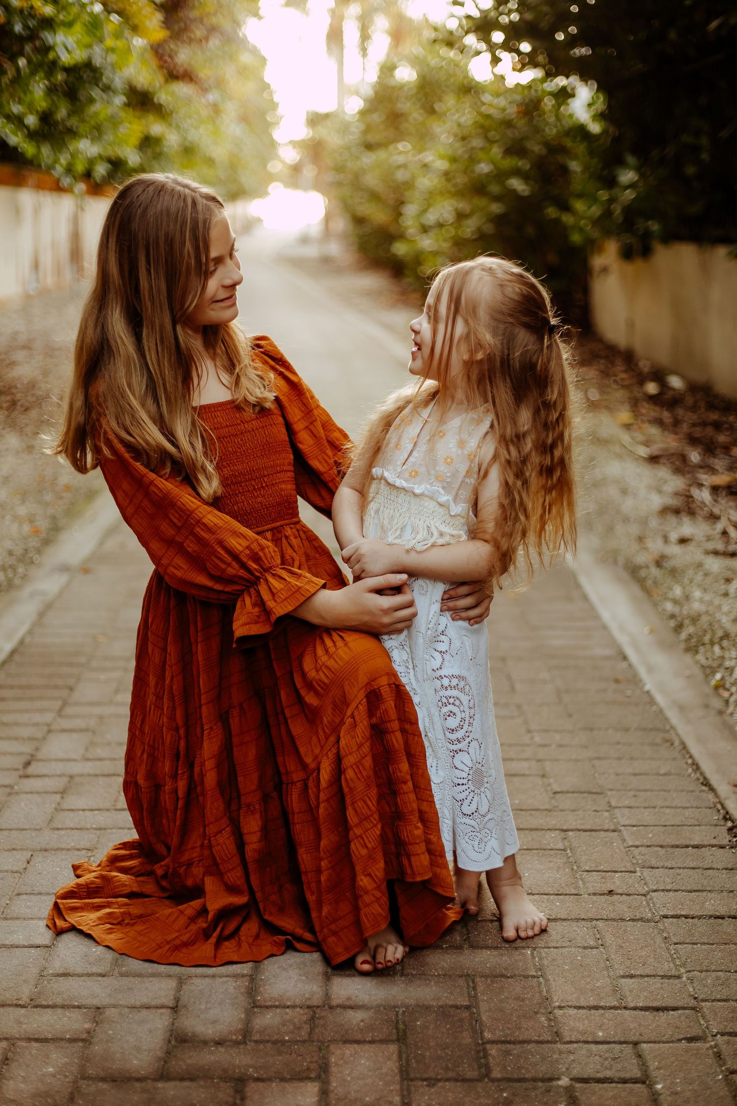 A tender moment between two people in flowing dresses, one rust-colored and one white, standing on a brick pathway at sunset.