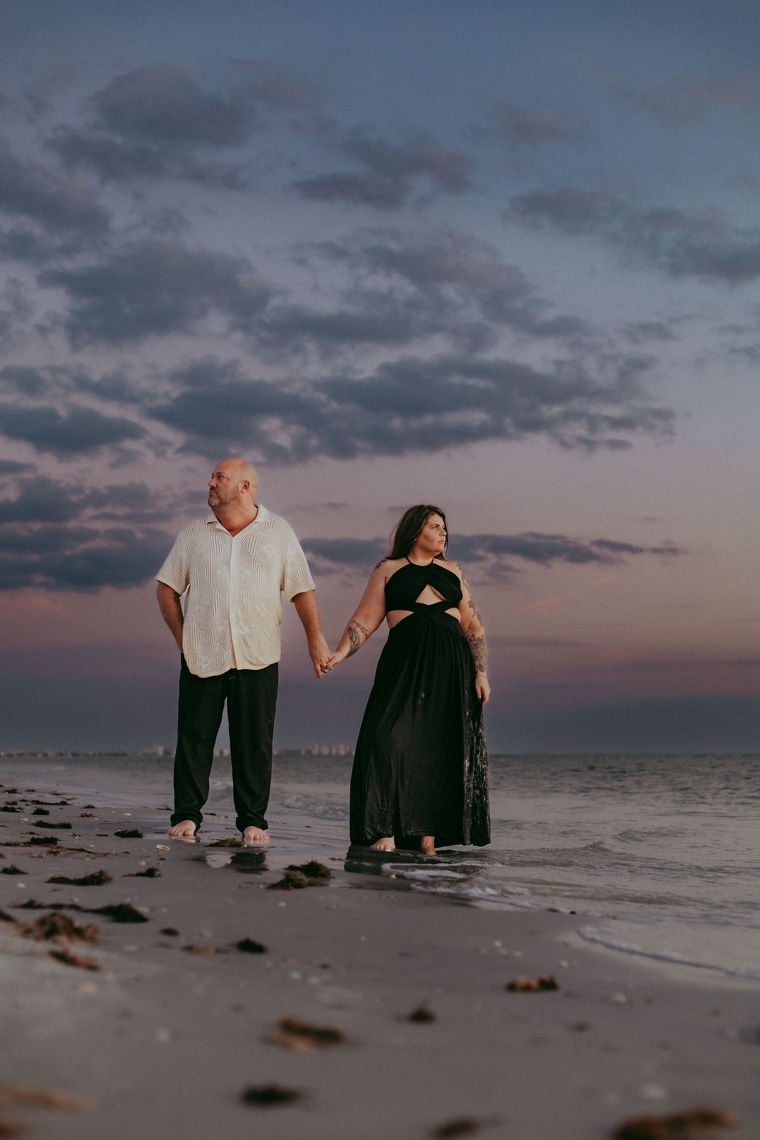 A couple holds hands while walking along a beach at sunset with dramatic cloudy skies overhead.