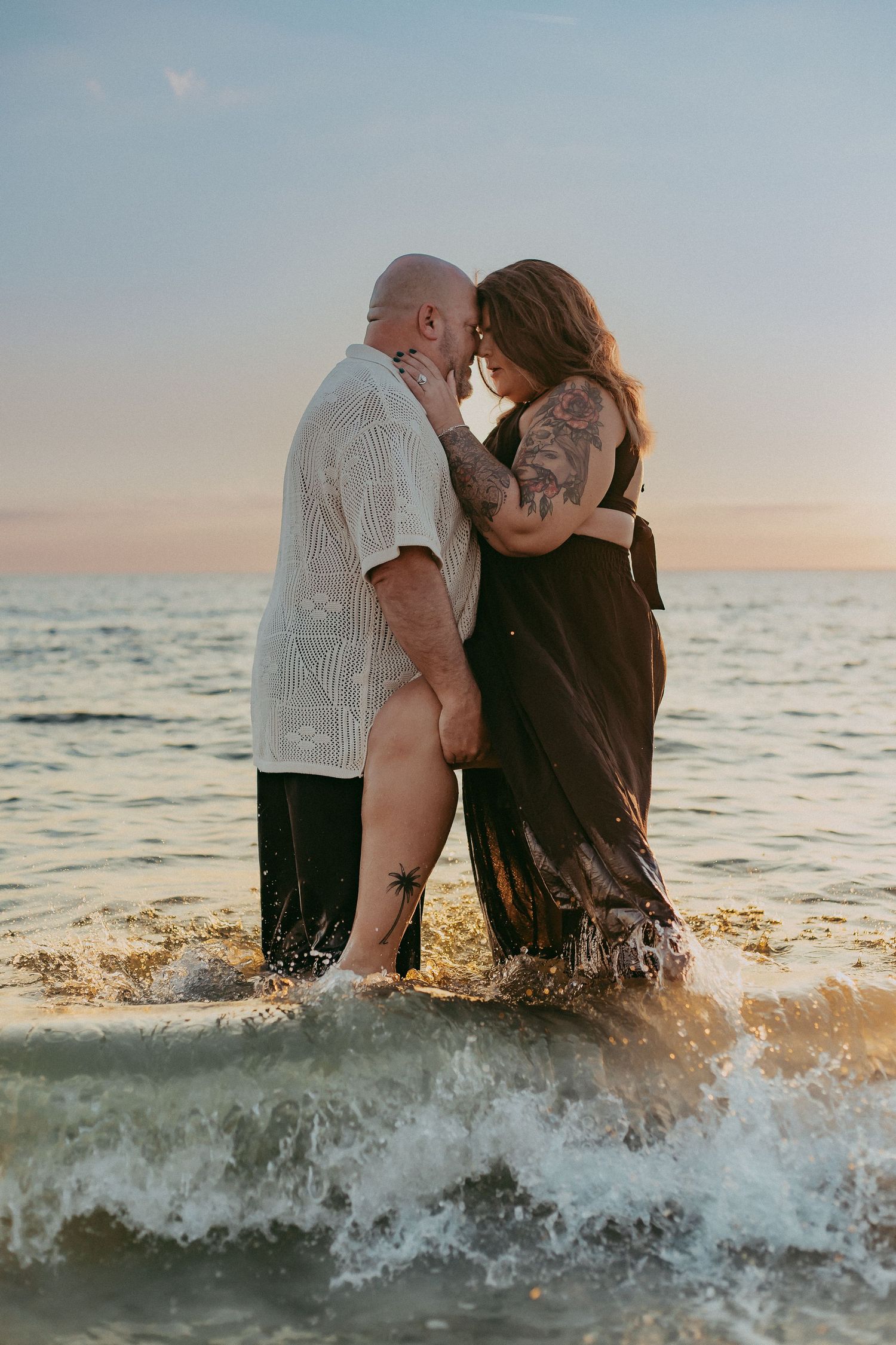 A romantic couple embraces in the ocean waves at sunset during a beach photoshoot.
