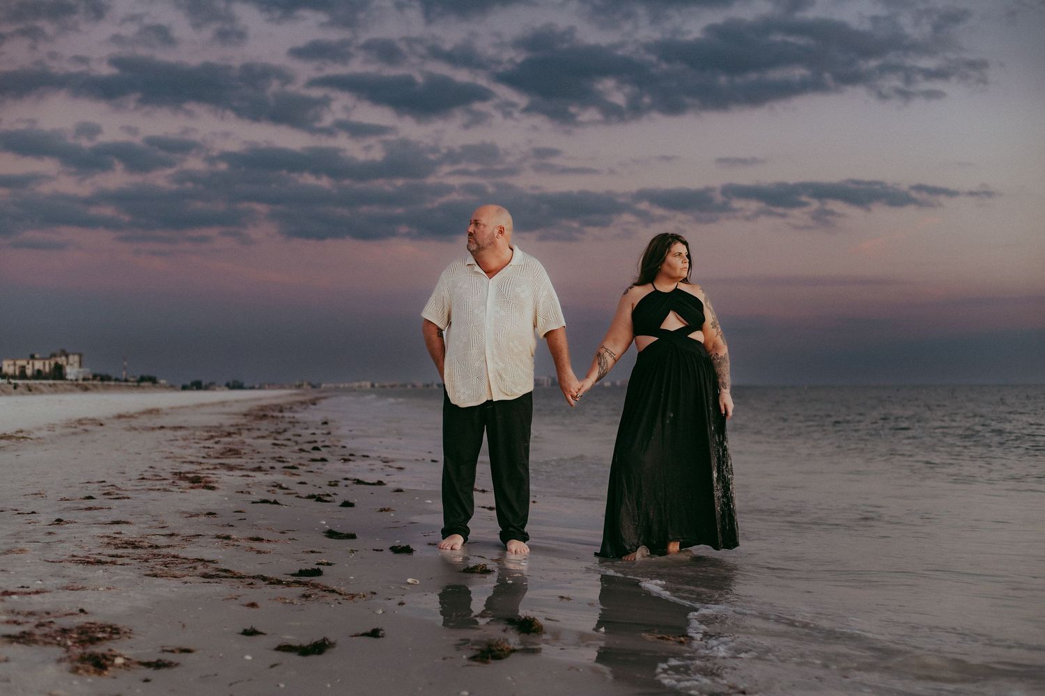 A couple walks together on a sandy beach at sunset with dramatic cloudy skies overhead.