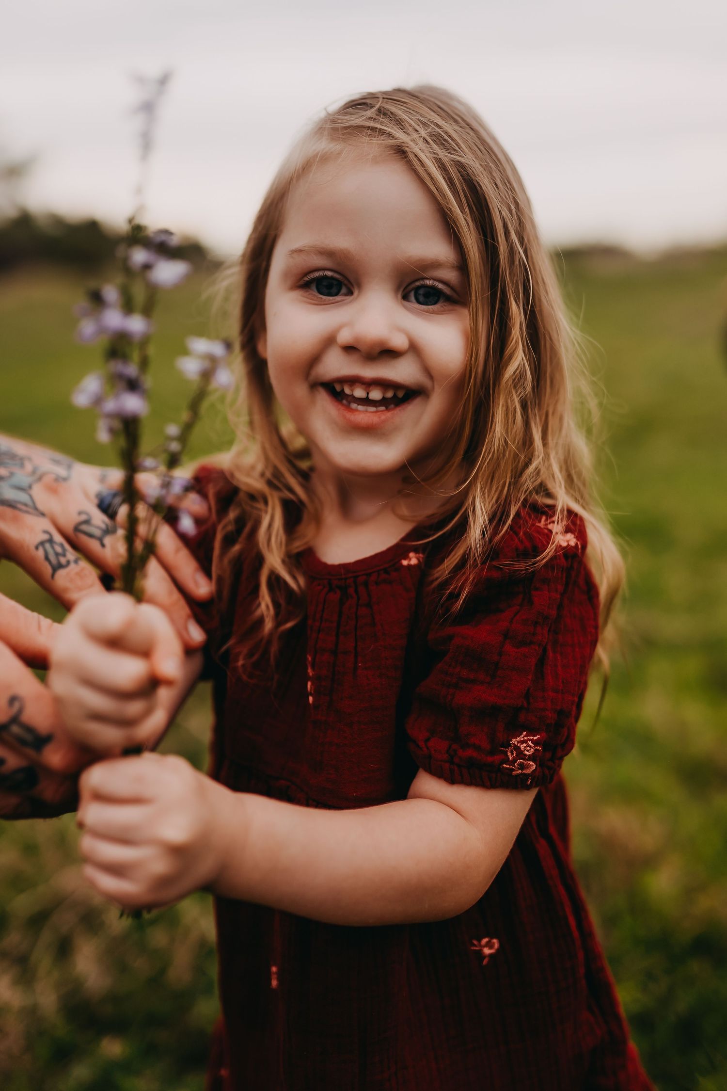 Child in burgundy dress holds purple wildflowers in grassy field at sunset.