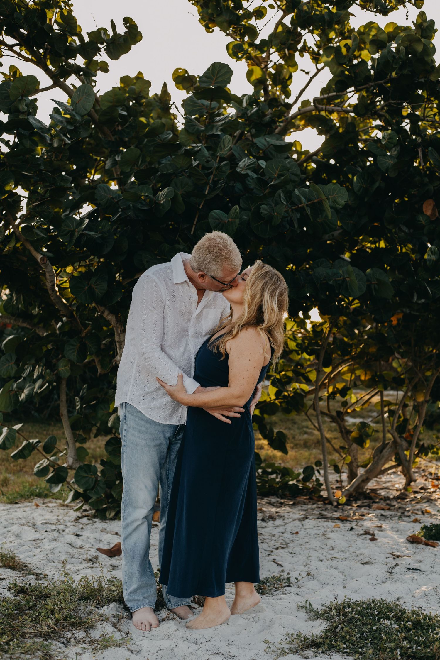 A romantic couple embraces on a sandy beach under lush green foliage during golden hour.
