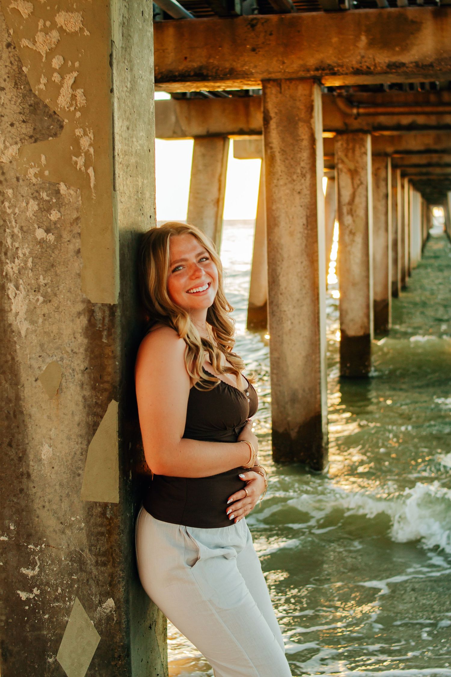 A person laughs joyfully while posing under a pier with waves and sunlight streaming through the wooden pillars.