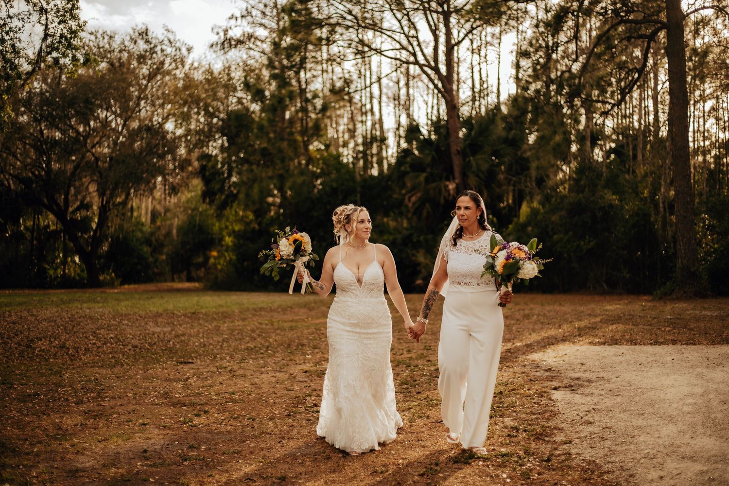 Two people in elegant white wedding dresses hold hands while walking on a dirt path at sunset in a wooded area.