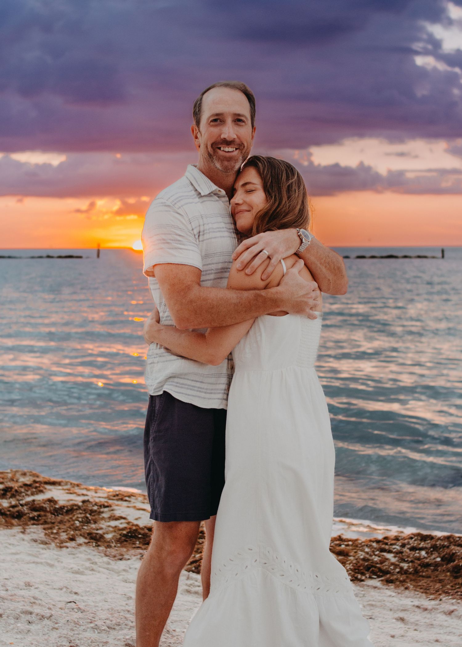 A couple embraces on a beach at sunset with dramatic purple clouds and orange sky over the ocean.