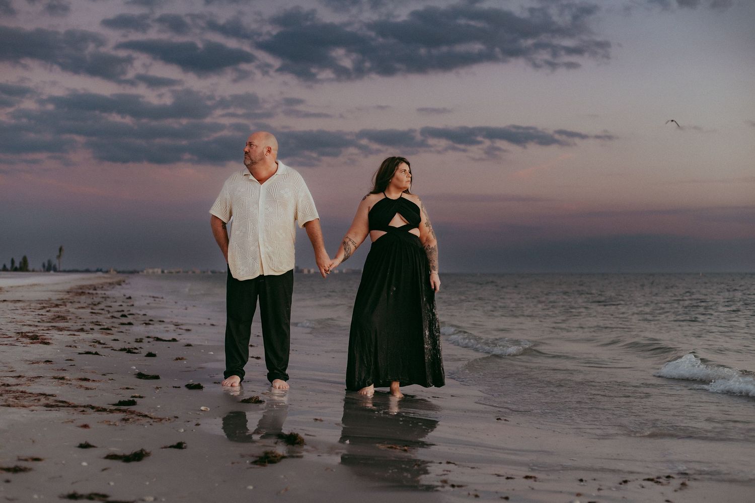 A couple walks hand in hand along a beach at sunset with dramatic clouds and ocean waves in the background.