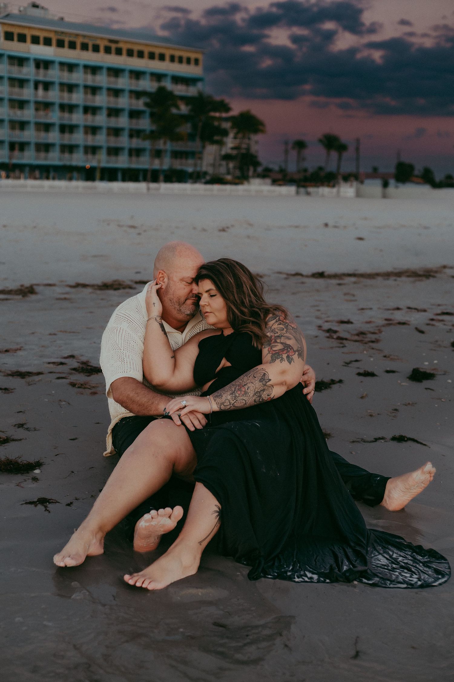 A couple embraces on the beach at sunset with a hotel and palm trees in the background.