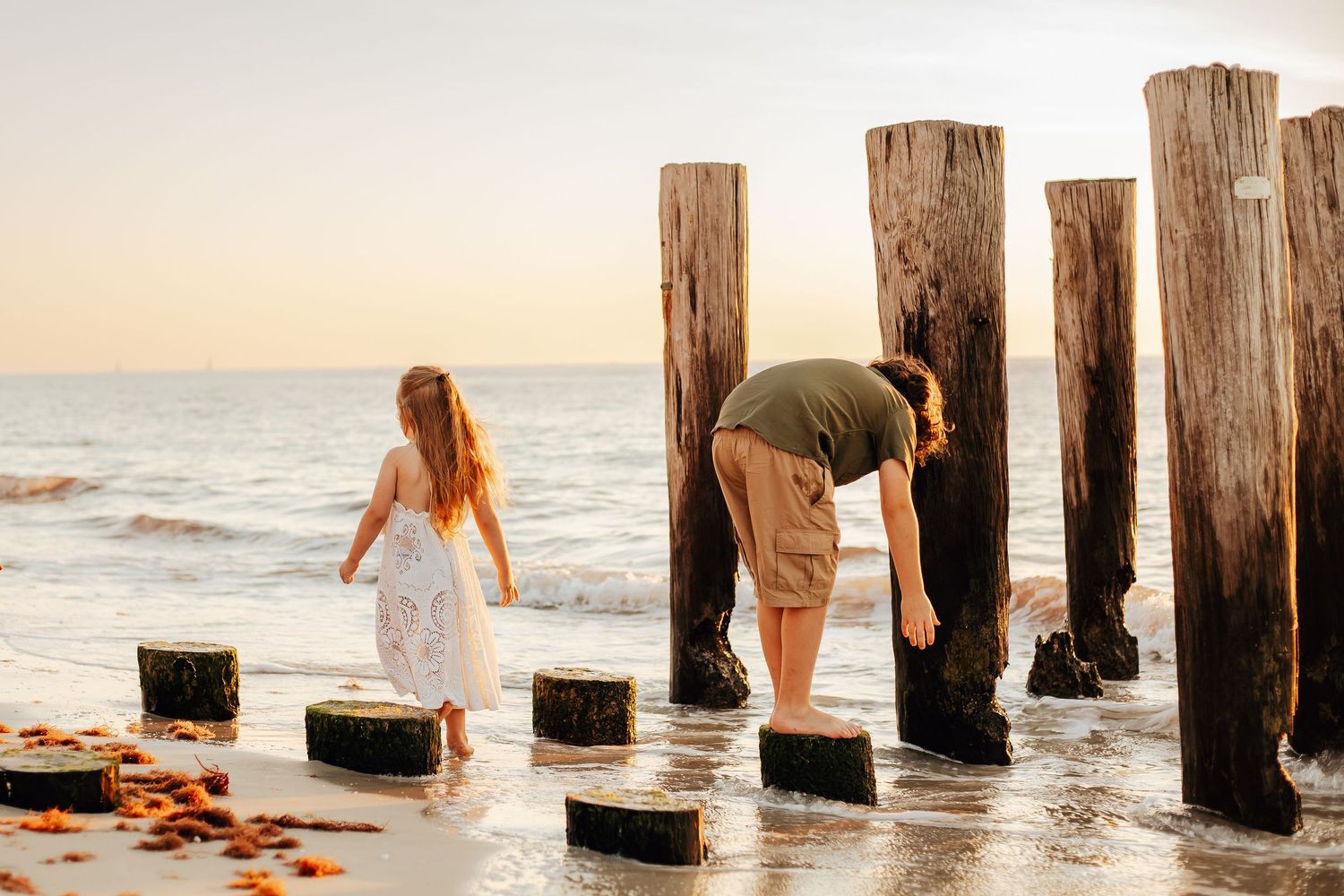 A person in a white dress walks along wooden beach pilings during sunset at the ocean's edge.