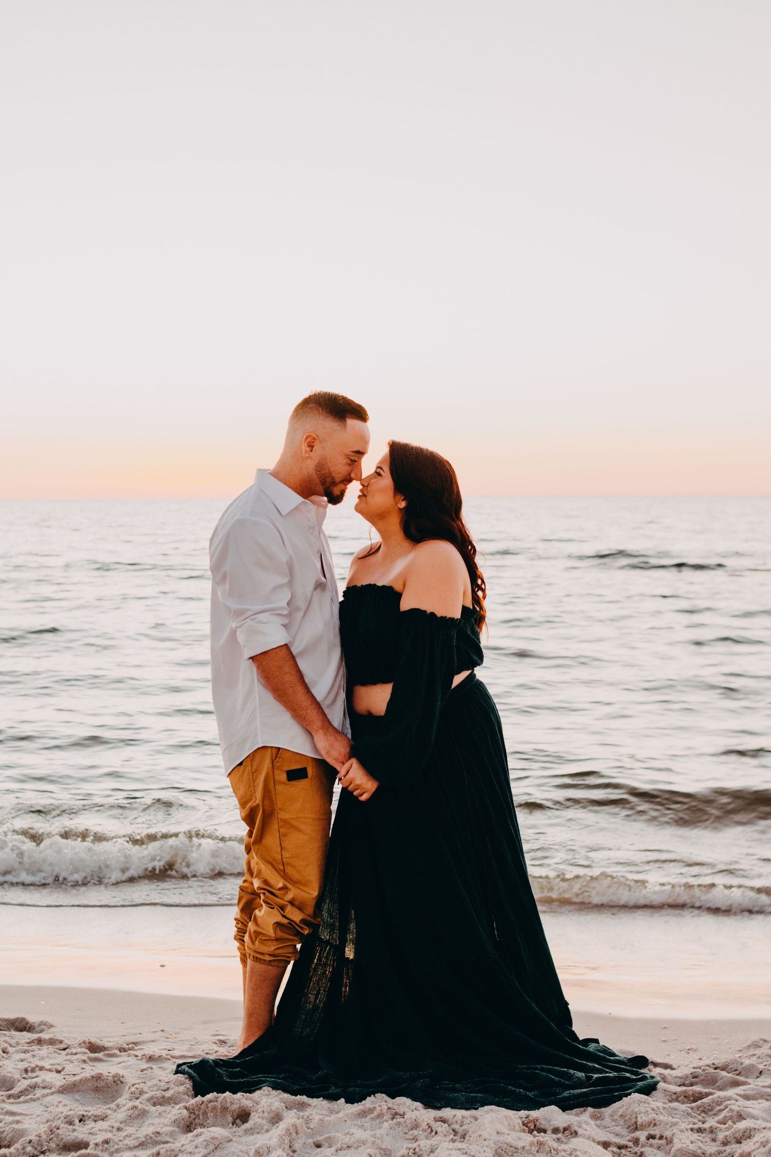 A couple shares a romantic moment at sunset on a sandy beach with waves rolling in the background.