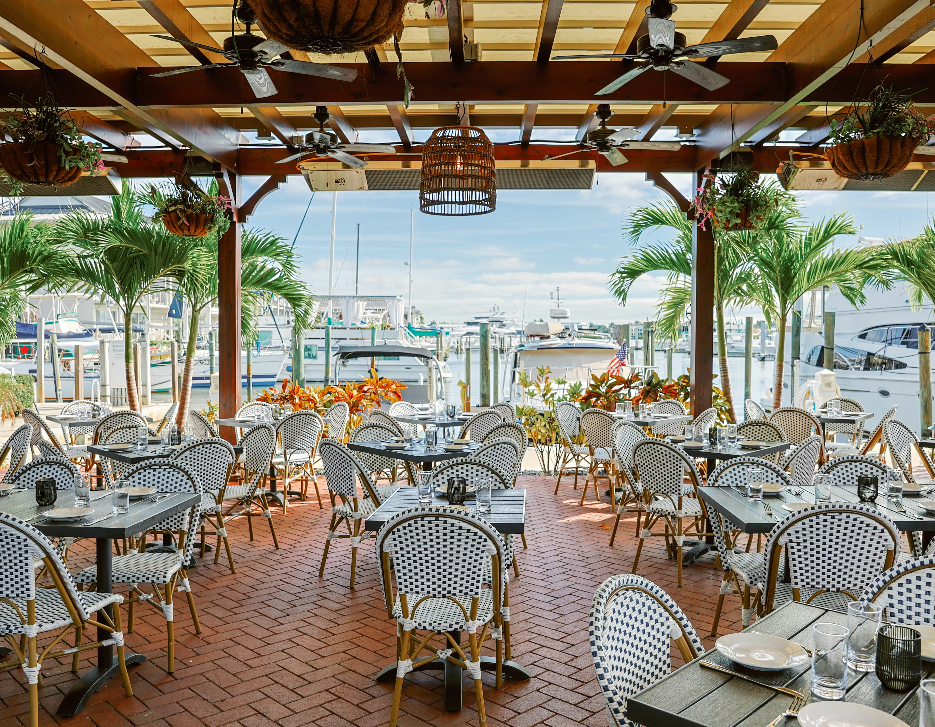 Waterfront restaurant patio with striped chairs and tables overlooking a marina with boats and palm trees in the background.