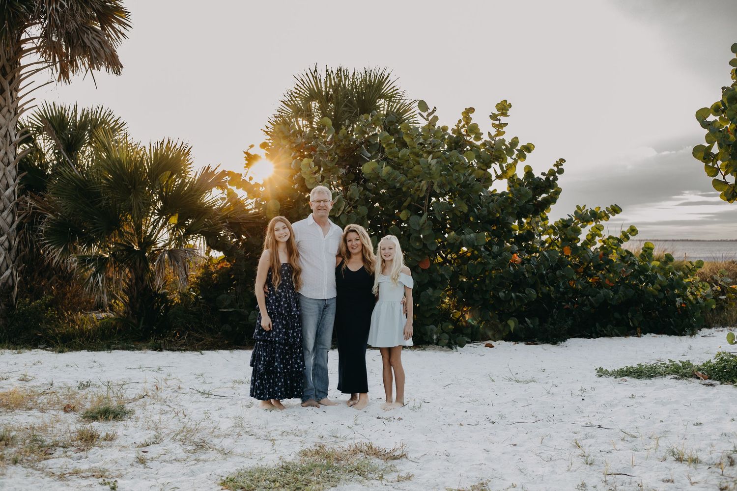 Three people stand together on a sandy beach at sunset with palm trees and greenery in the background.