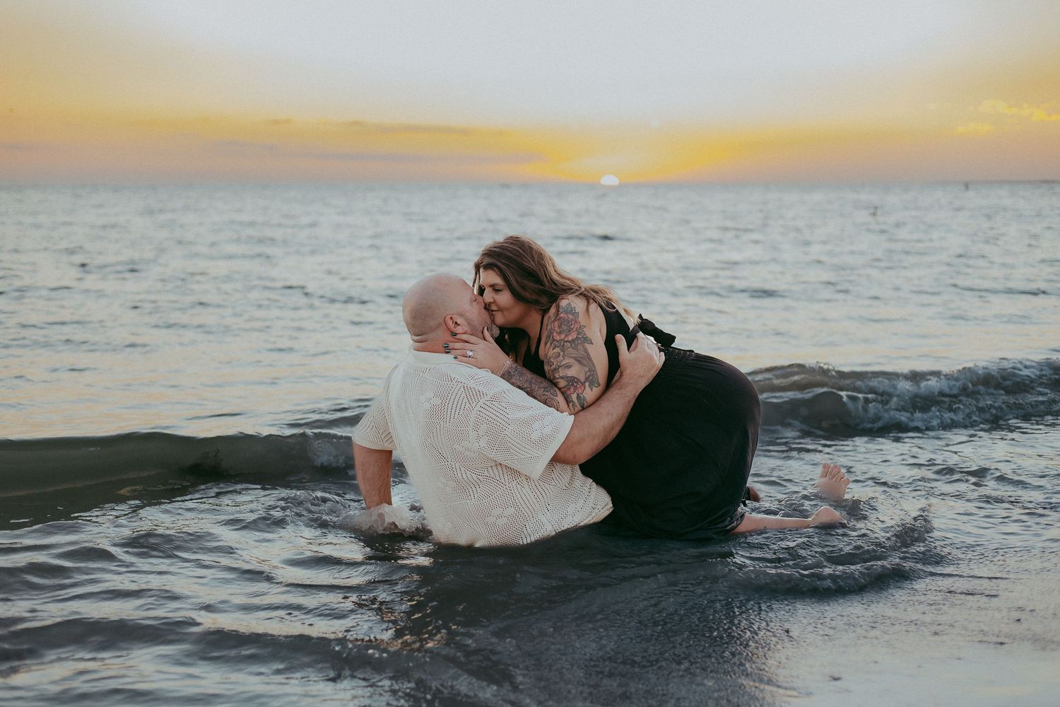 A couple sits on rocks by the ocean during a romantic sunset photoshoot at the beach.