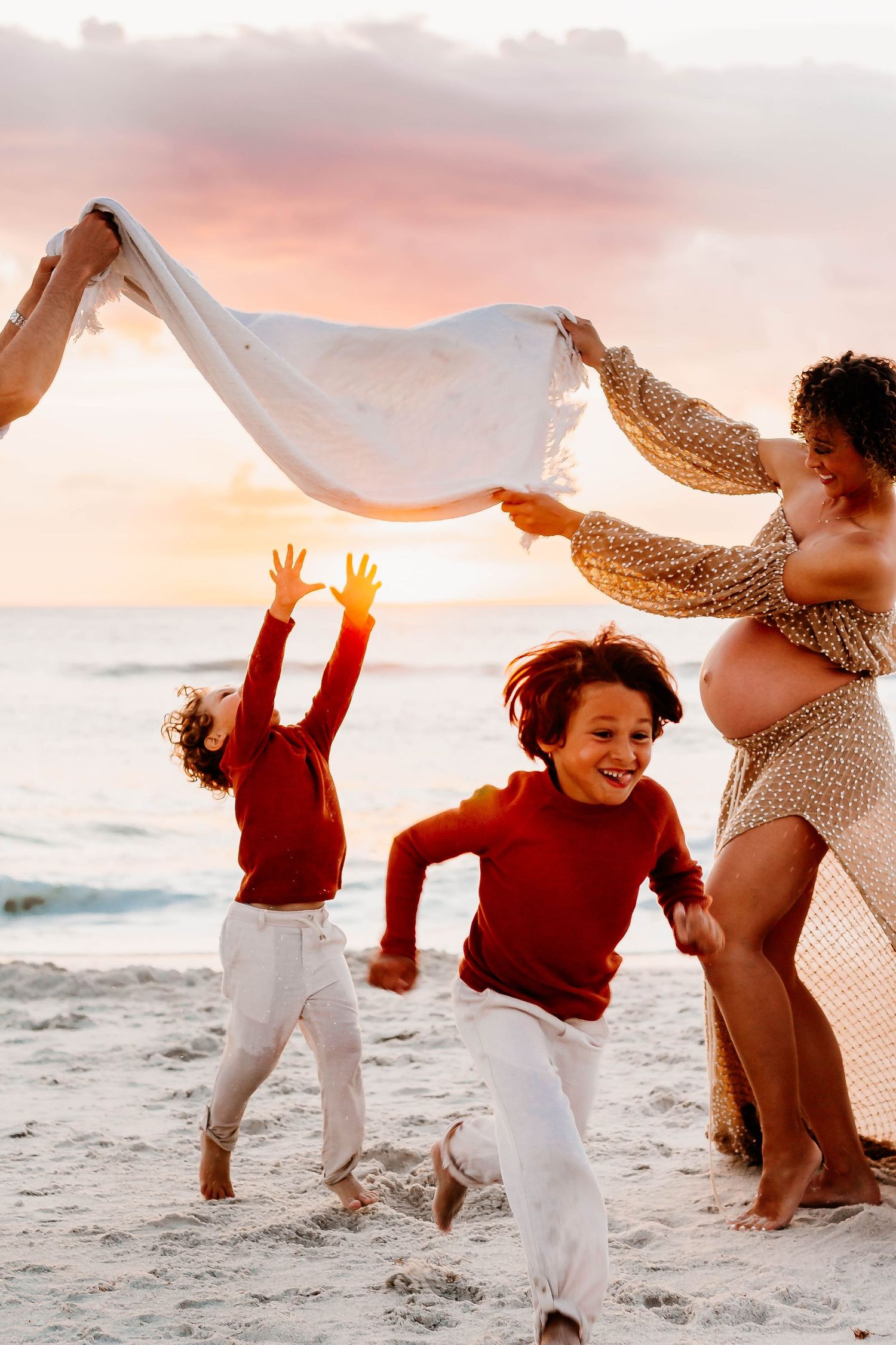 Family playing with white fabric on beach at sunset as waves crash along shoreline in joyful moment.