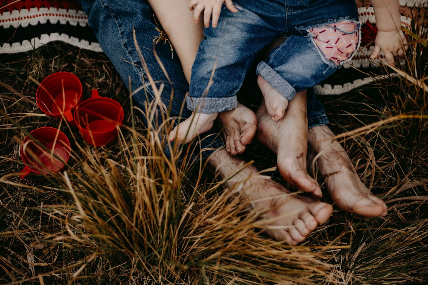Bare feet of adults and child resting on dry grass with red tropical flowers in a family outdoor photo.