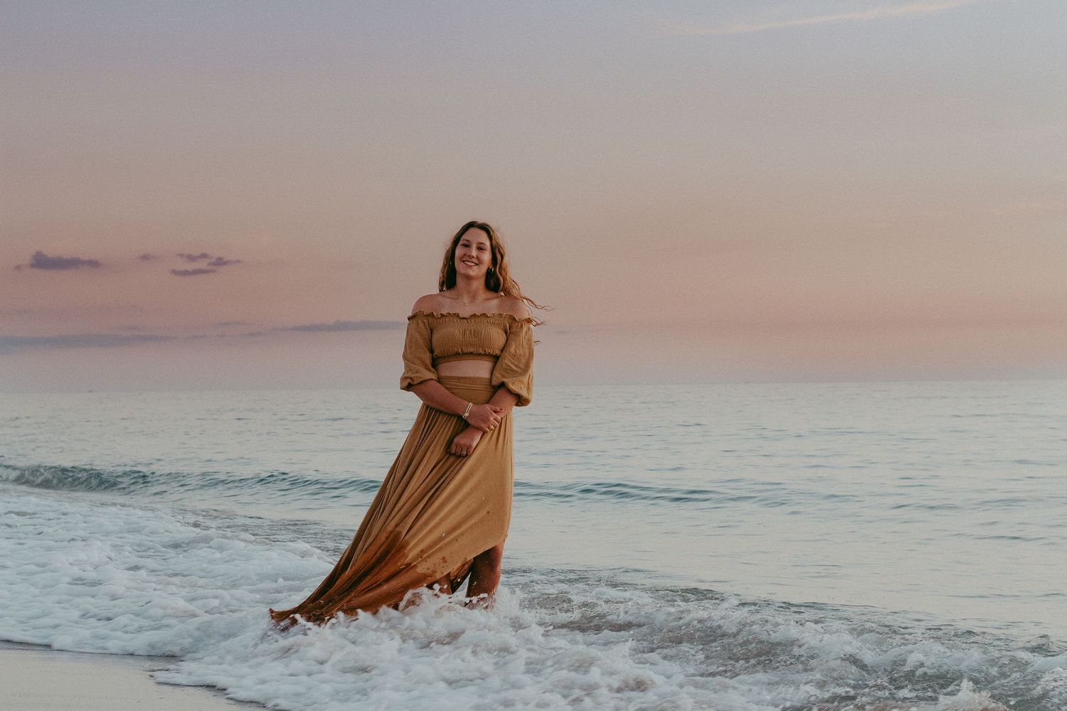 A person in a flowing beige dress stands in ocean waves during a pink sunset at the beach.