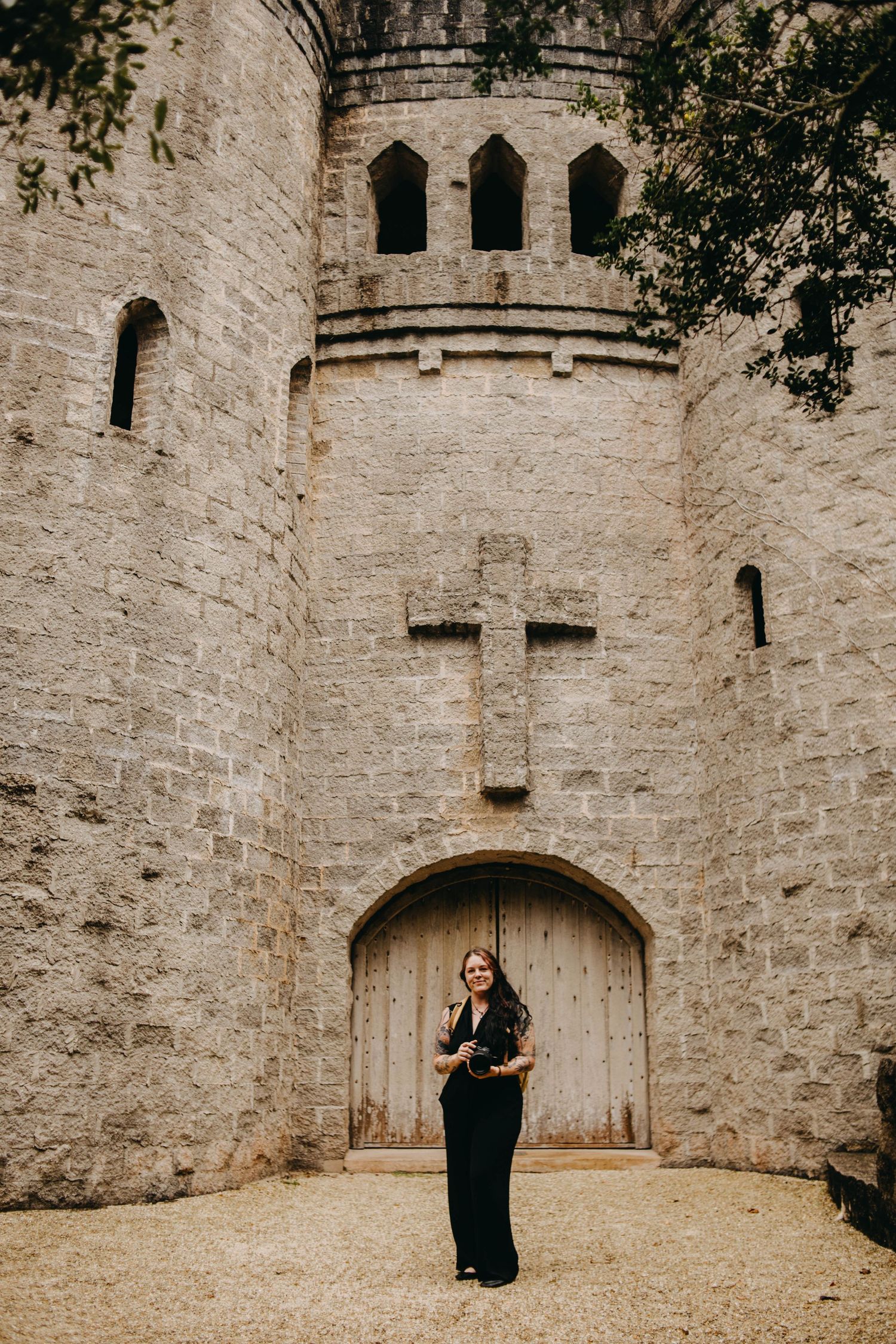 Woman in black outfit standing before a stone castle entrance with a cross above wooden doors.