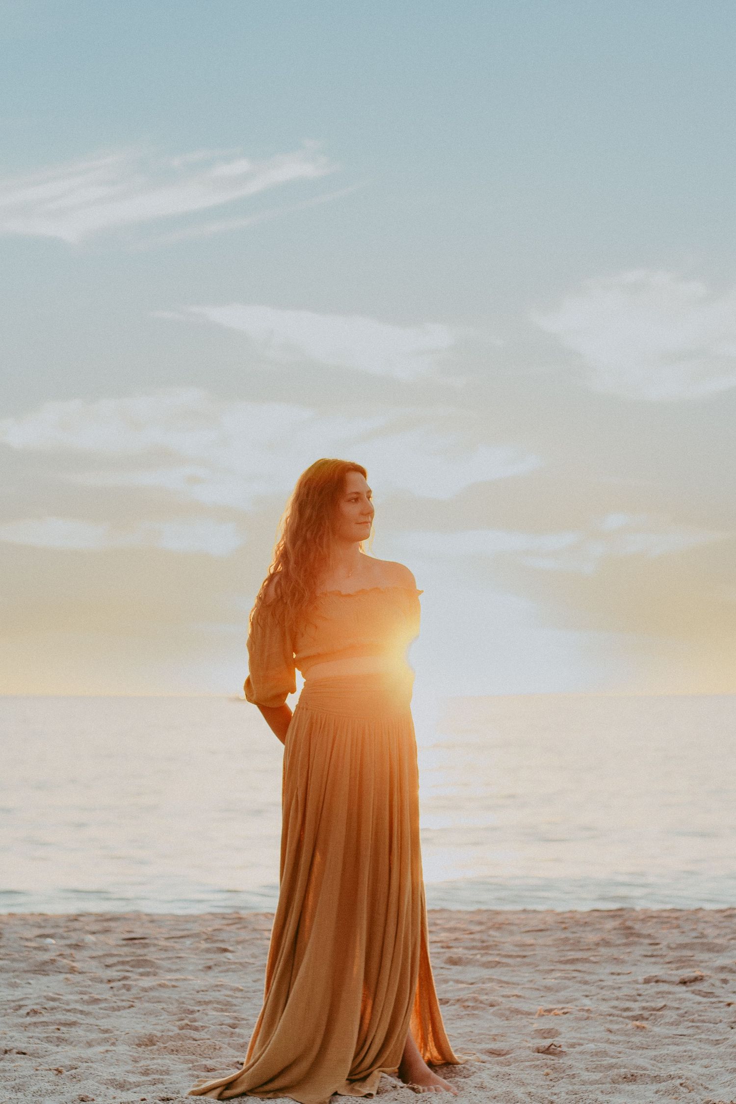 A figure in a flowing dress stands on the beach at sunset with golden light streaming through their silhouette.