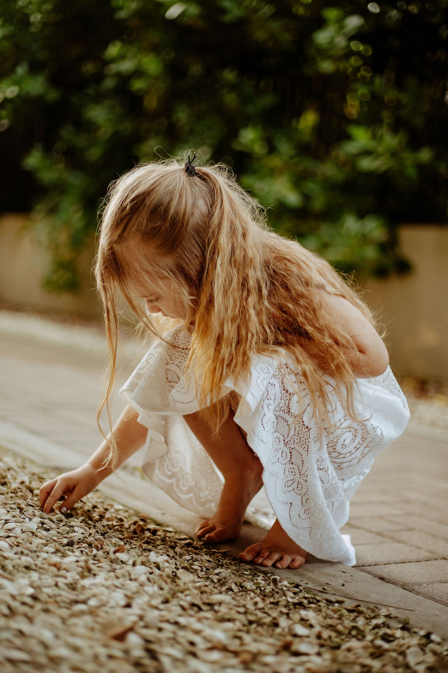 A person in a white lace dress bends down to examine something on a stone path in a garden setting.