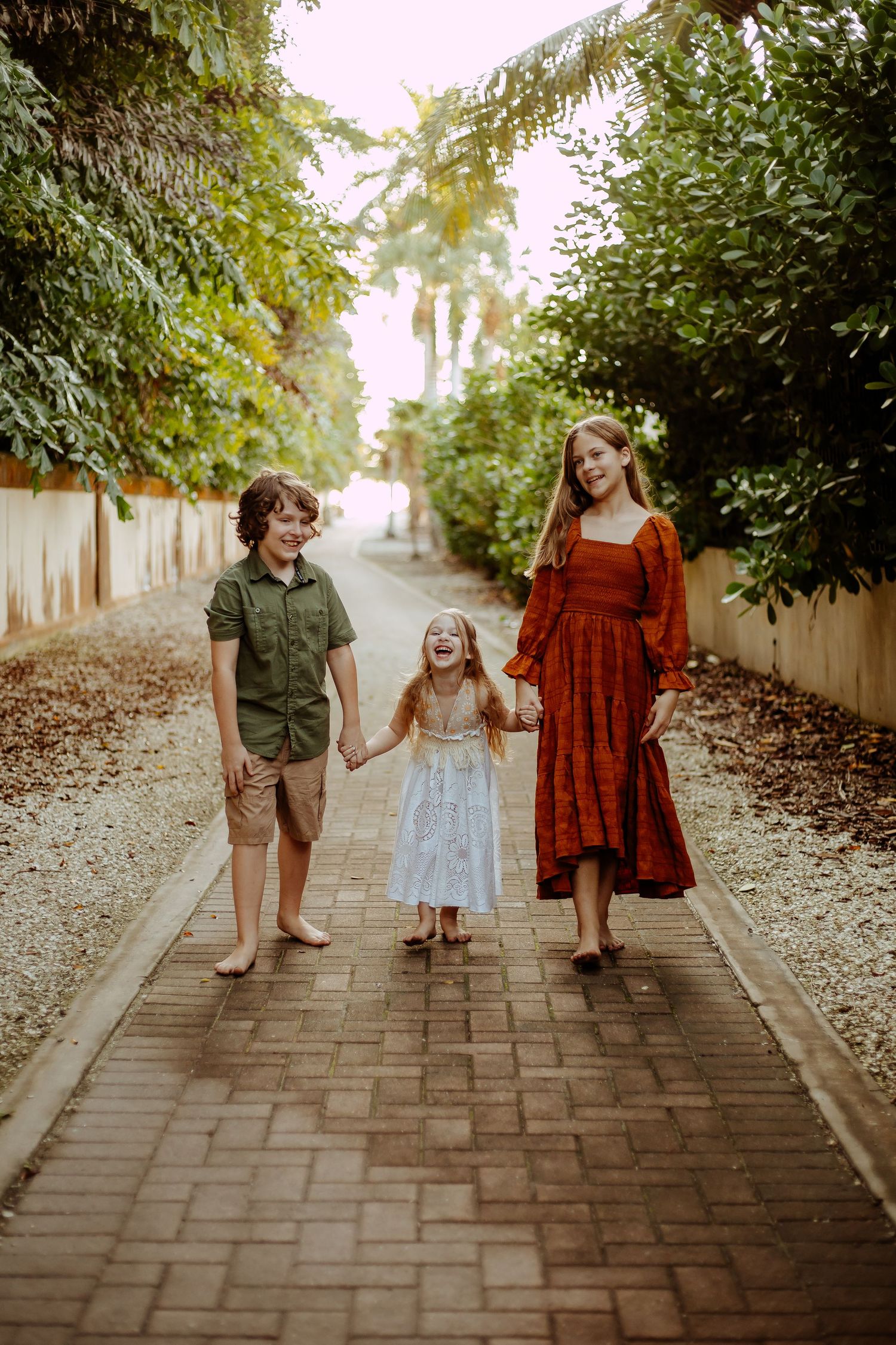 Three people walk and laugh together down a tree-lined brick pathway on a sunny afternoon.