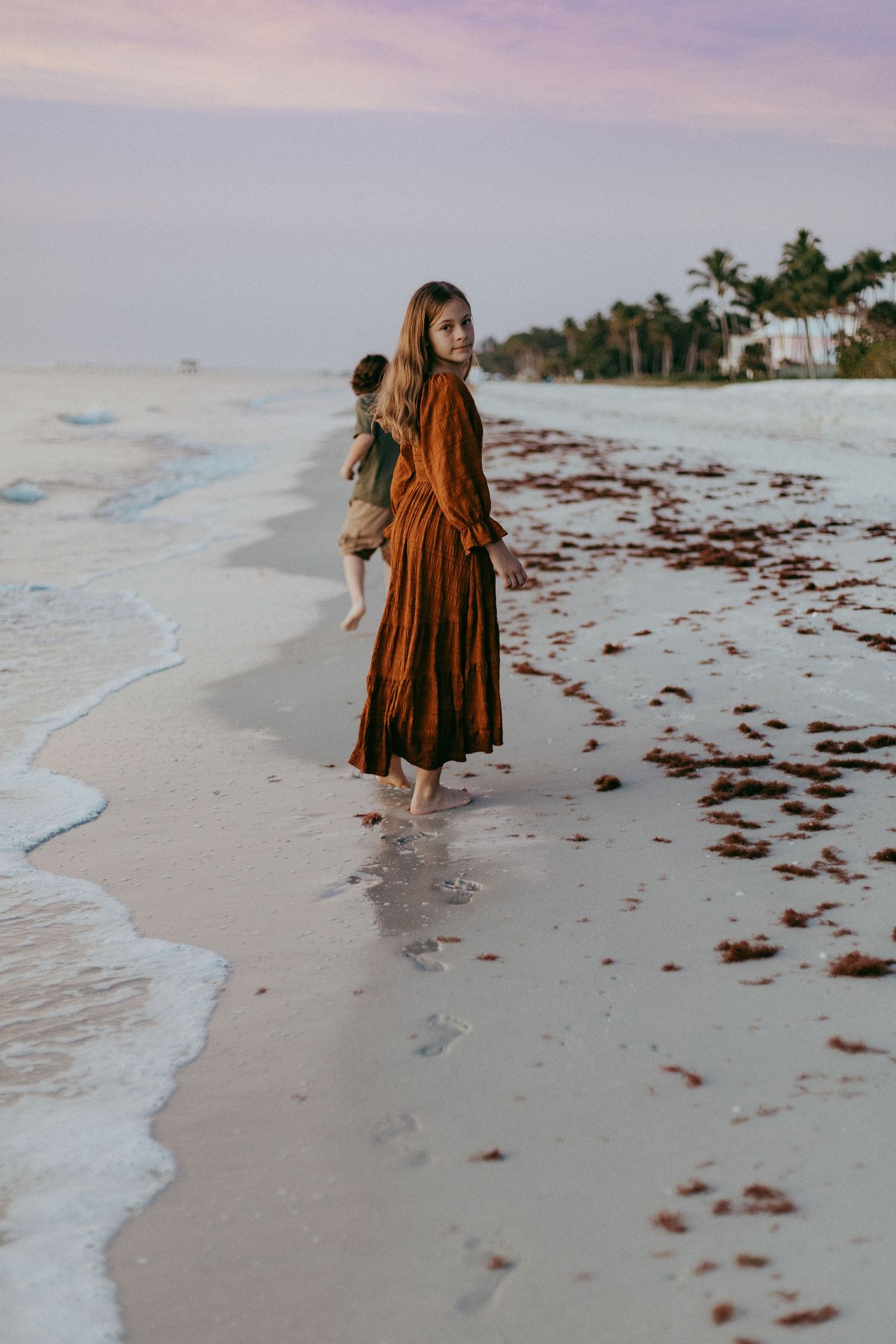 Person in a brown dress walks along a serene beach shoreline at sunset with seaweed scattered on white sand.