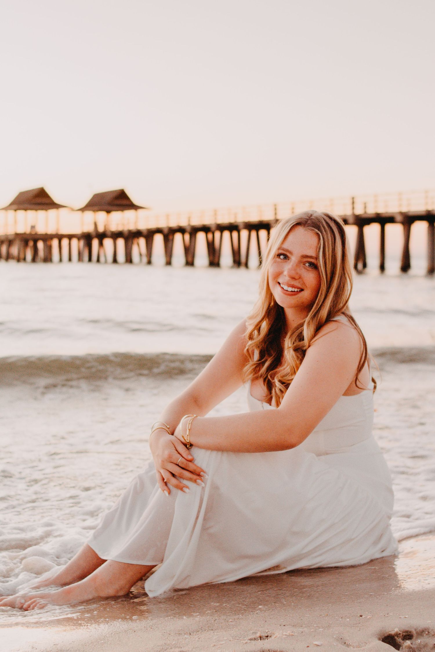 Woman in white dress lounging on beach at sunset with pier in background.