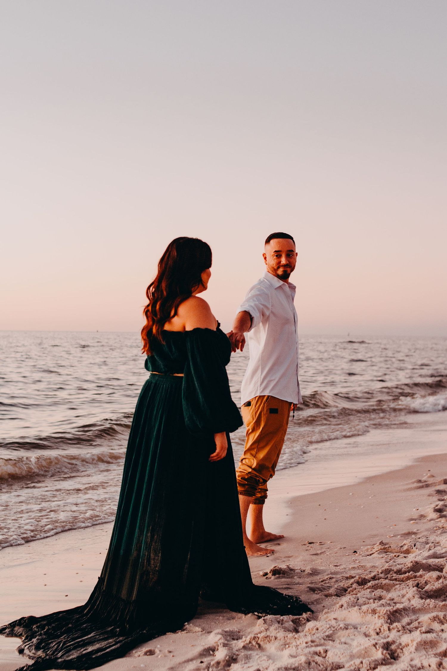 A couple stands together on a beach at sunset, with the woman wearing a flowing black dress and the man in casual attire.