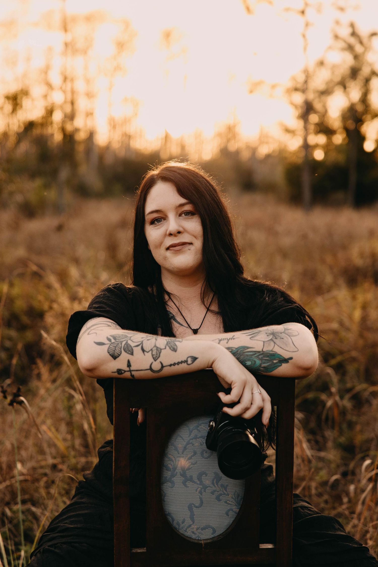 Woman with tattoos sits in a wooden chair in a golden field at sunset, holding a camera.