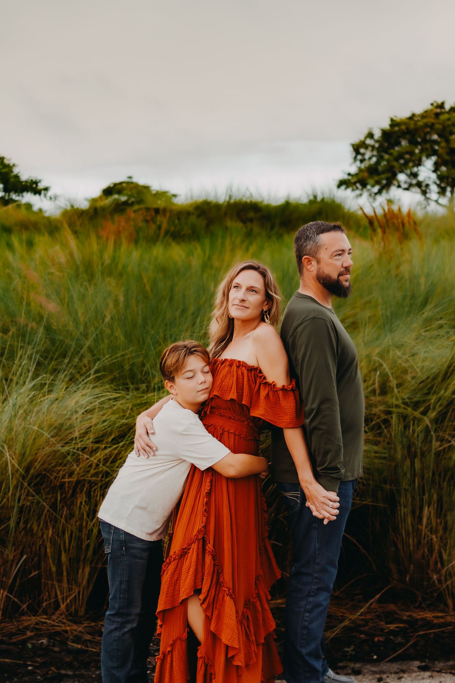 A family poses together in tall grass at sunset with a woman in a flowing red dress between two others.