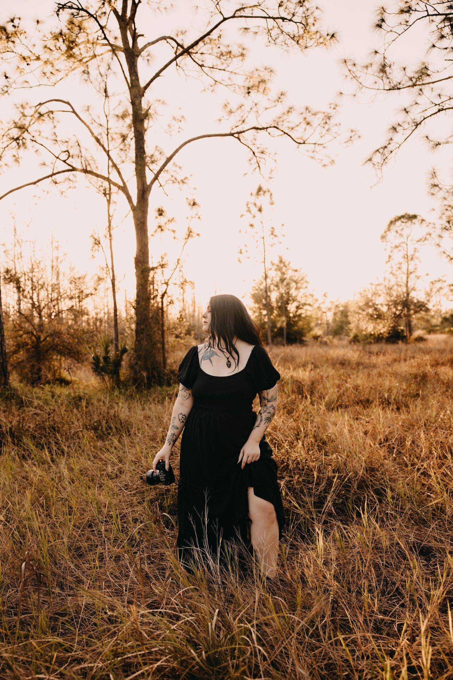 Woman in black dress with tattoos walks through golden field at sunset near bare trees.