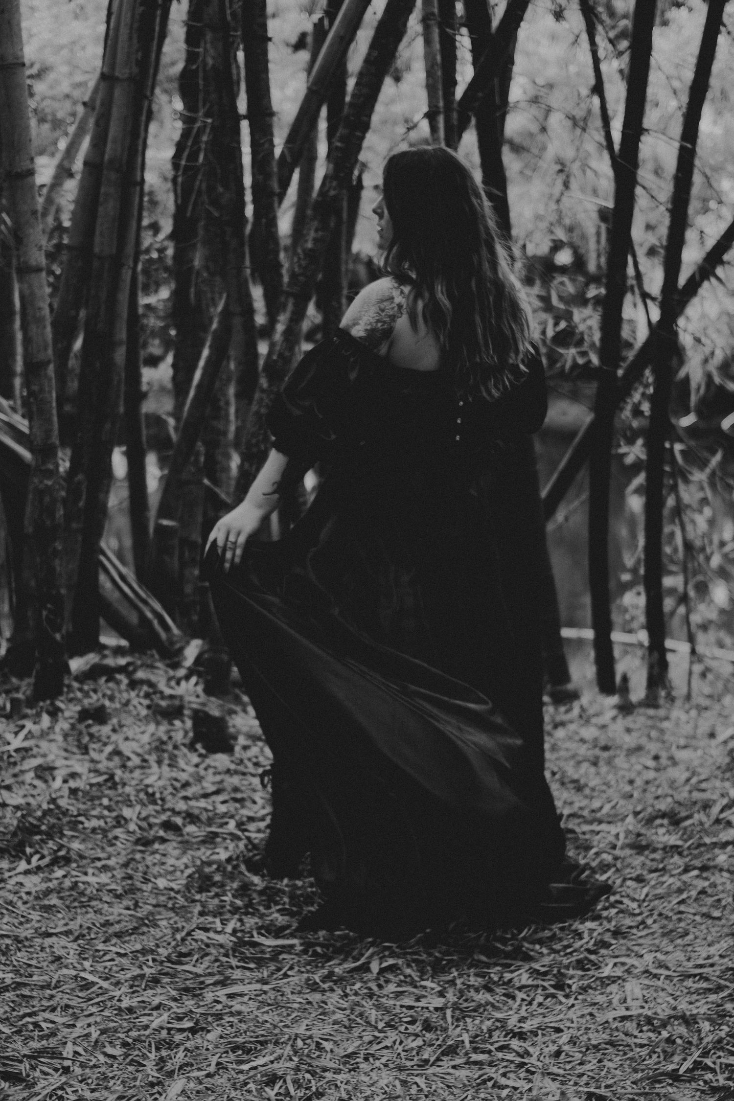 Black and white photo of a woman in a flowing dark gown standing among bare trees in a moody forest setting.