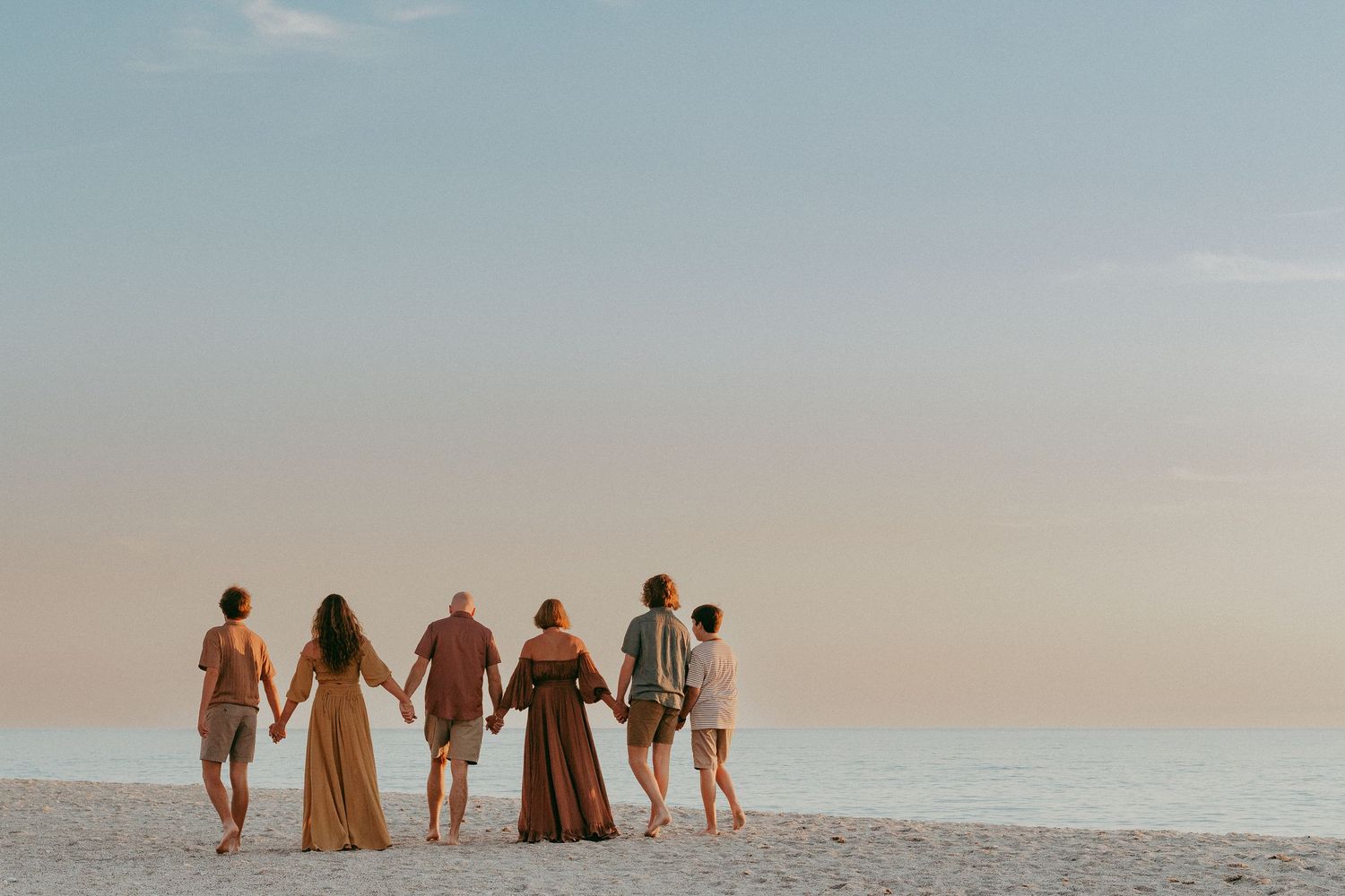 Group of friends holding hands while walking on a beach at sunset, creating silhouettes against the pastel sky.