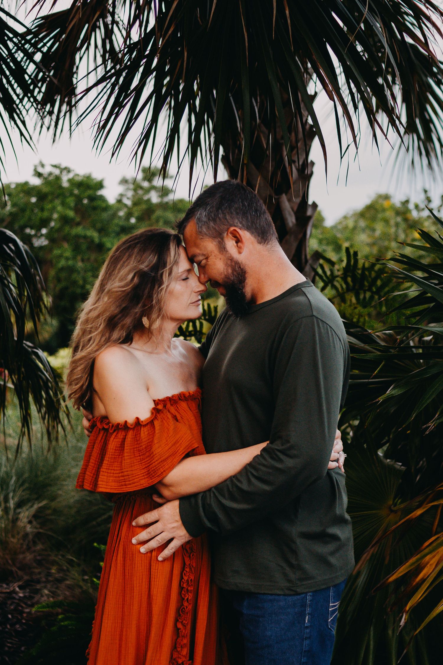 Romantic couple embraces in a lush tropical garden setting with palm trees at sunset.
