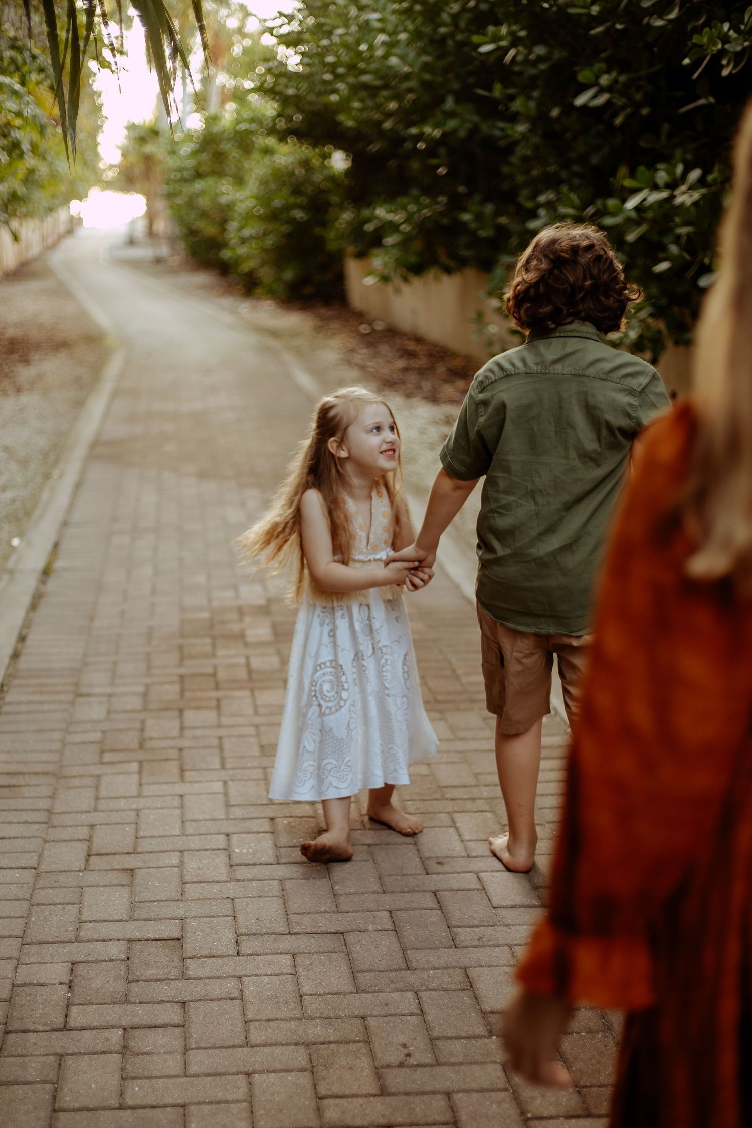 A child in a white dress walks with an adult down a brick-paved pathway surrounded by greenery.