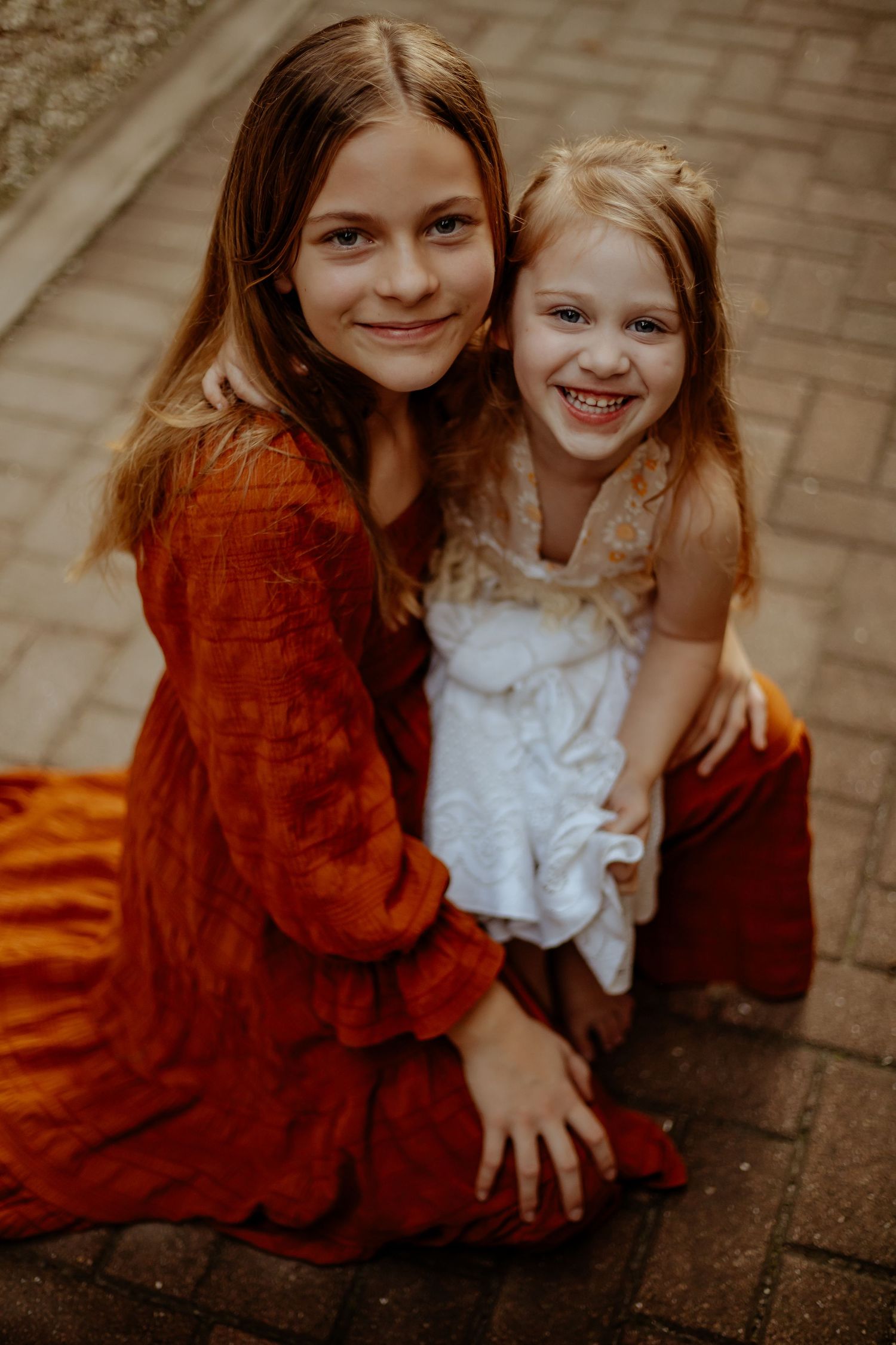 Two children sit close together on brick pavement, wearing contrasting rust-colored and white outfits.