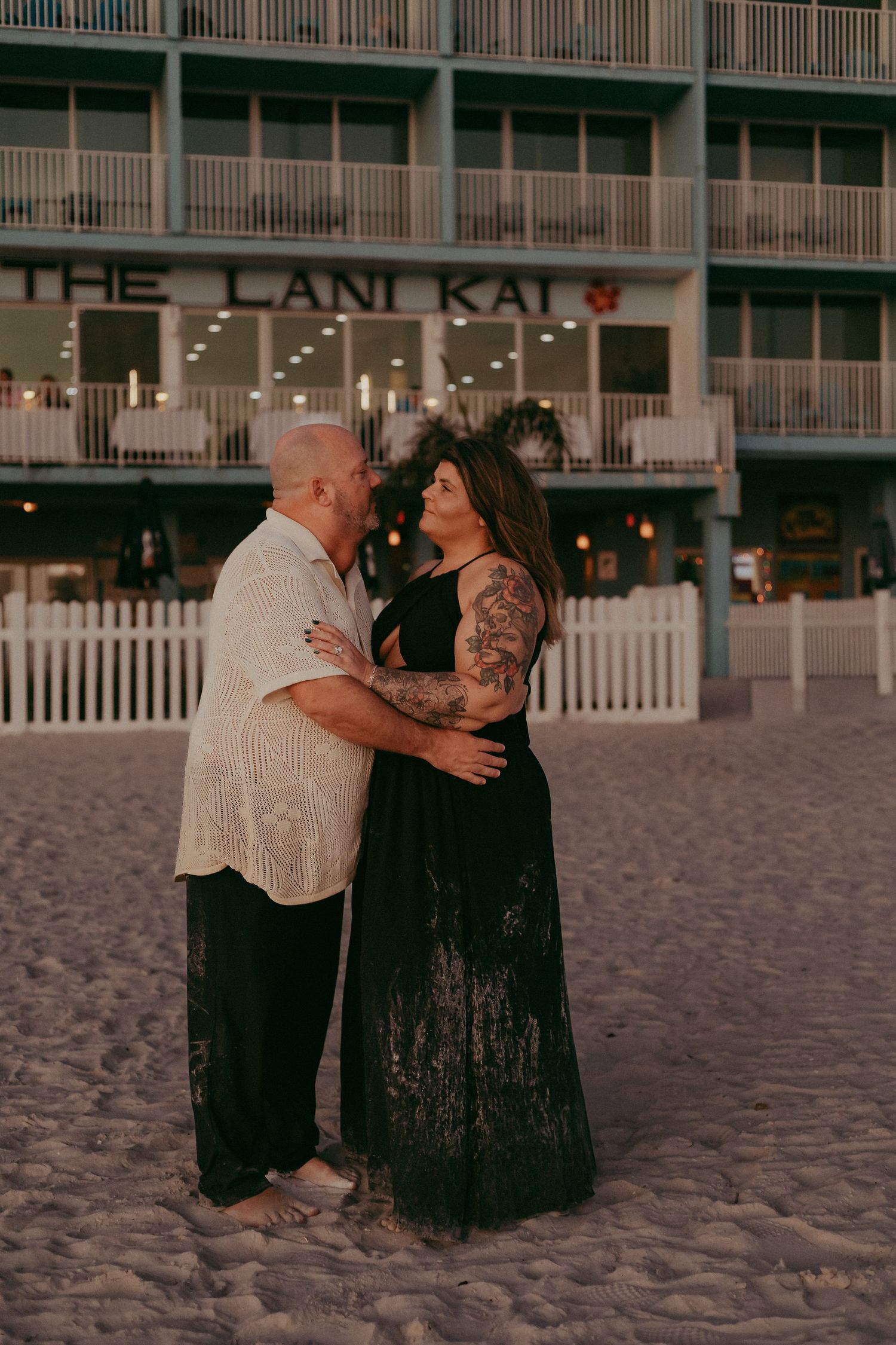 A couple dances romantically together on a sandy beach at sunset with a hotel building visible in the background.