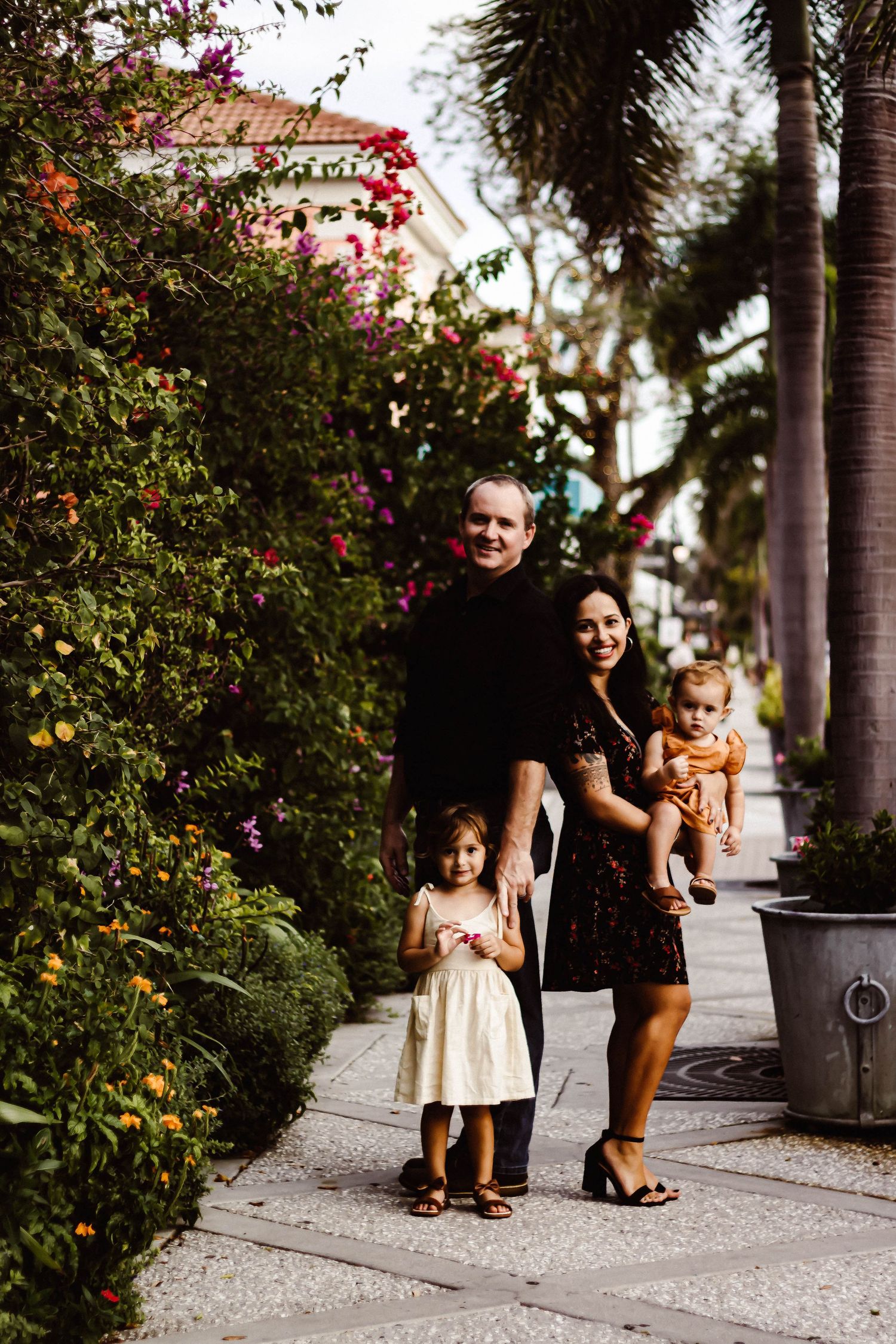 A family walks together along a palm-lined sidewalk with flowering bushes on a sunny afternoon.