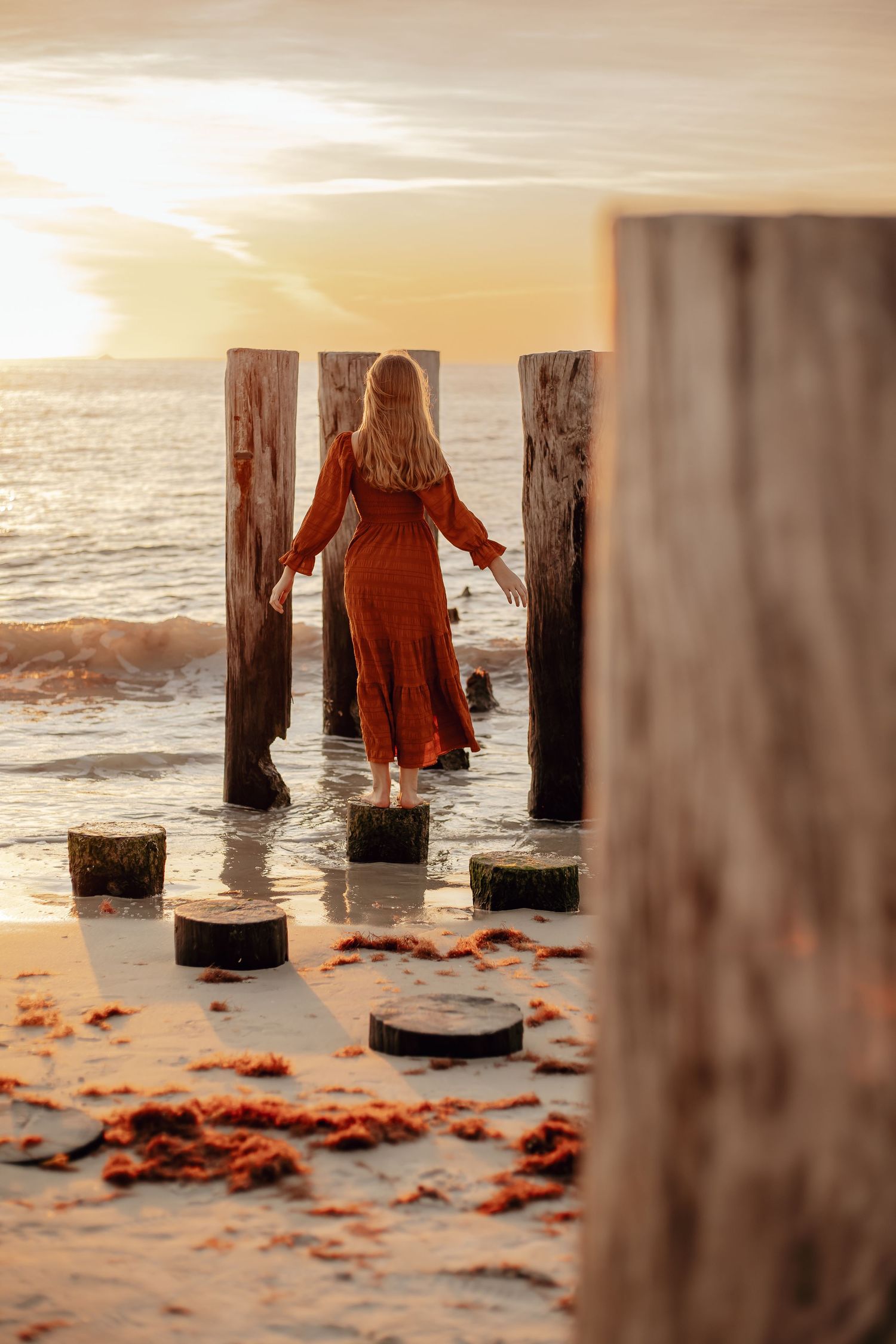 A red-coated figure walks among wooden posts on a sandy beach at sunset with scattered autumn leaves.