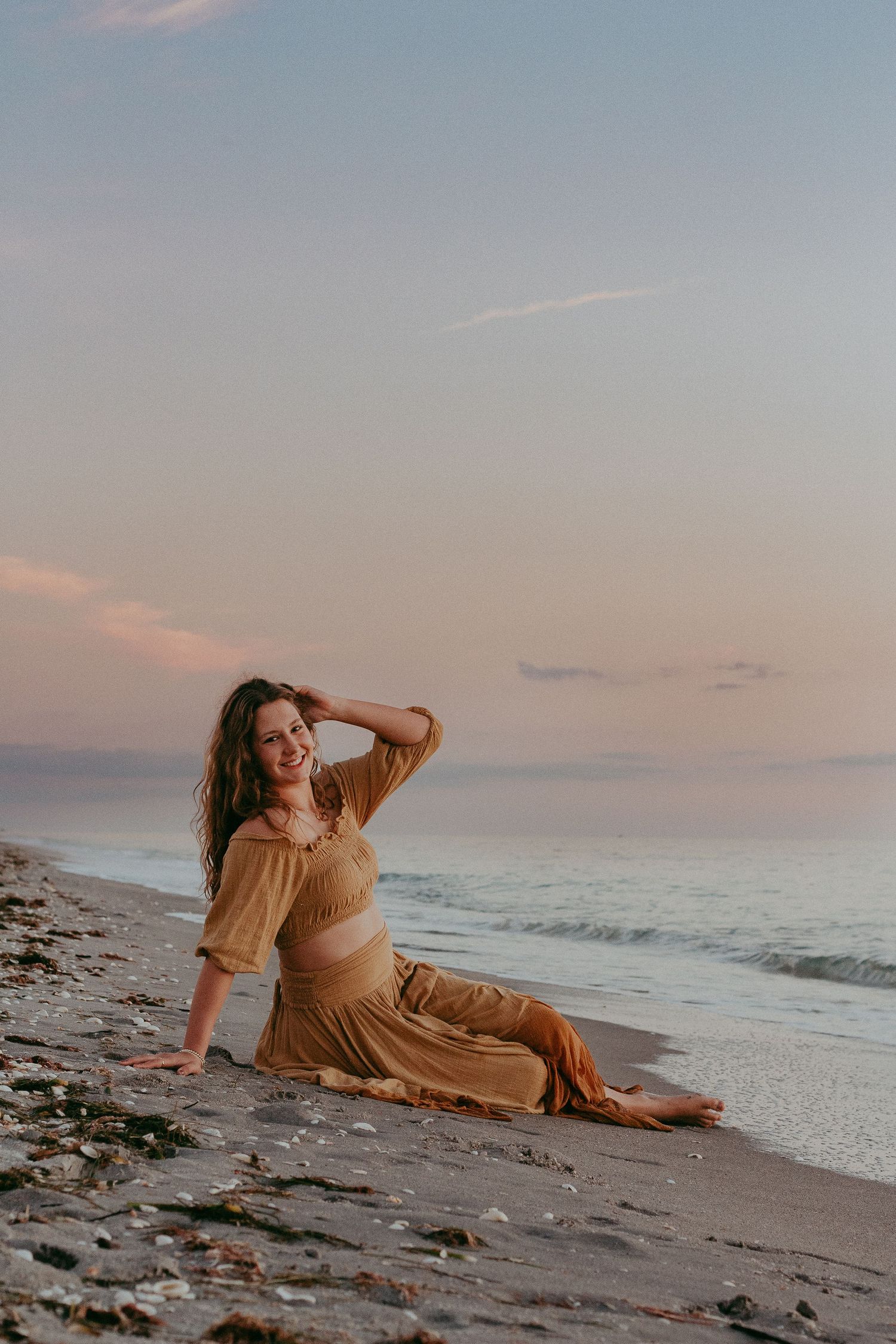 A person in a flowing tan dress relaxes on a sandy beach at sunset with ocean waves in the background.