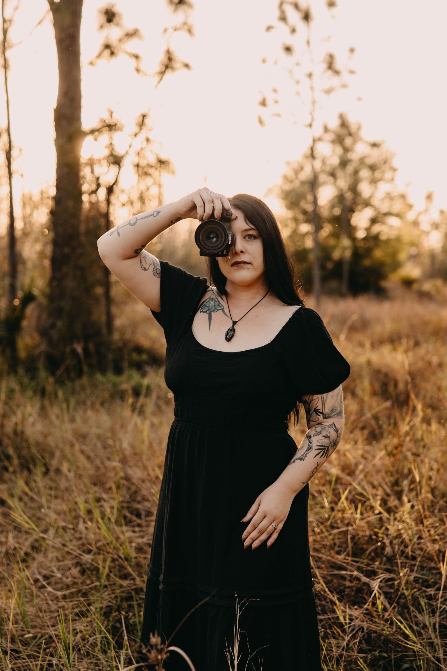 A person in a black dress poses expressively against a golden sunset in a field with tall grass and trees.